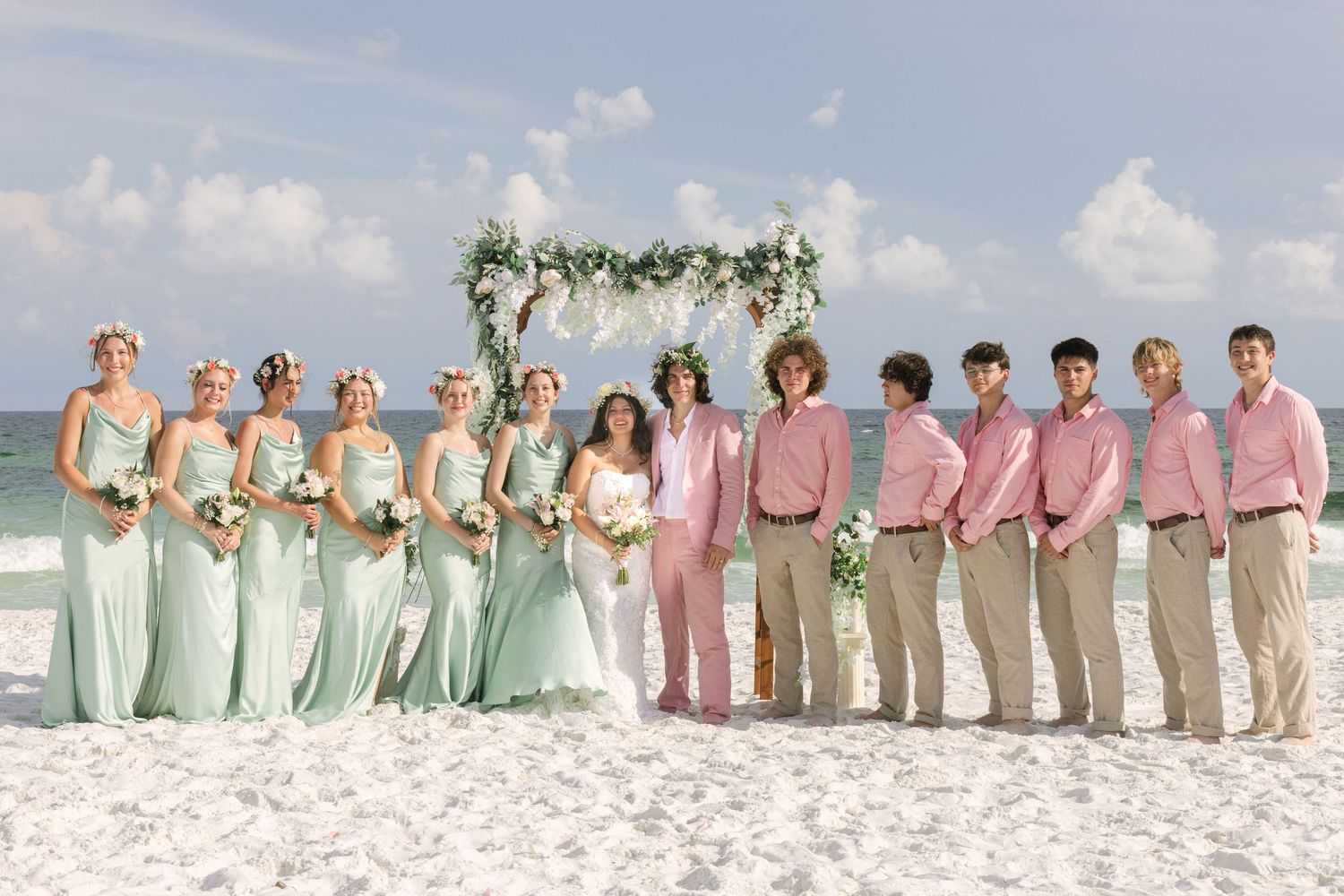 Beach wedding party group photo with coordinated pastel attire on white sand.