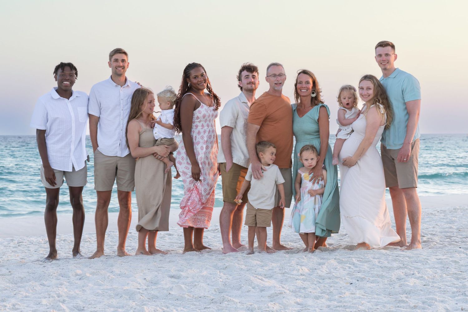 Large family group portrait on a sandy beach at sunset, wearing coordinated pastel outfits with ocean waves in the background.