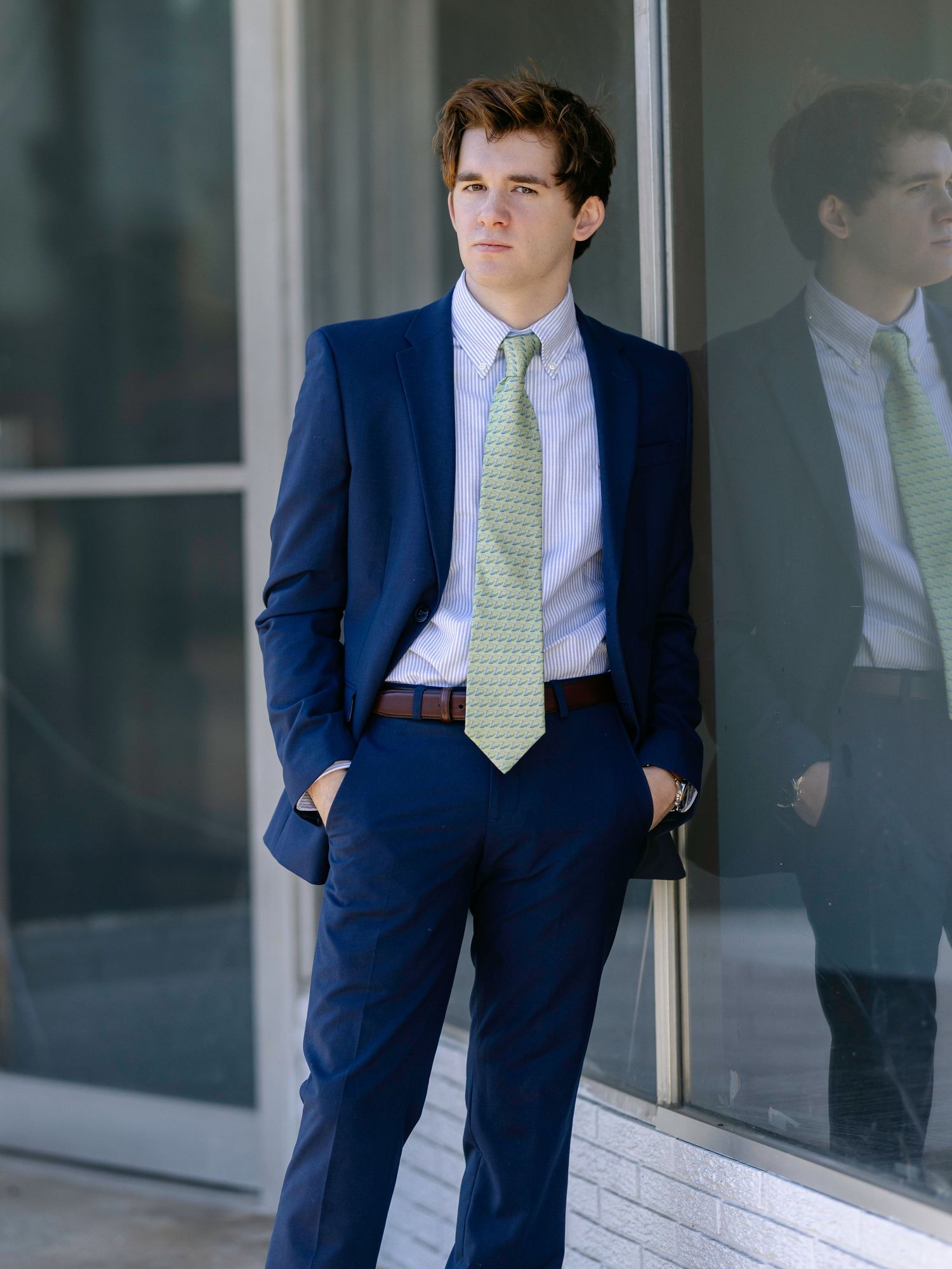 Young man in navy blue suit with green tie stands confidently outside modern building, hands in pockets.