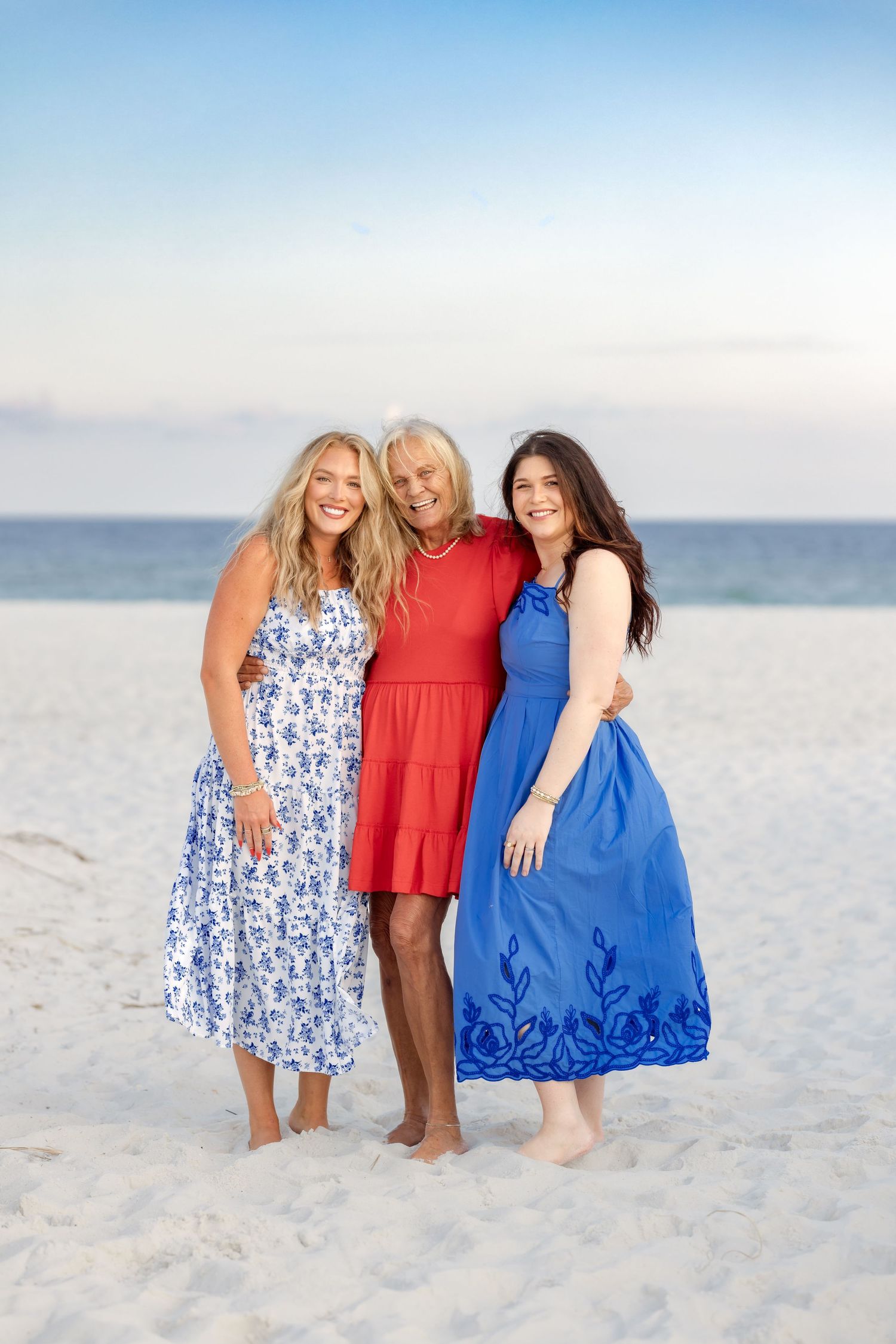 Three women in red, white, and blue dresses smiling together on a sunny white sand beach.