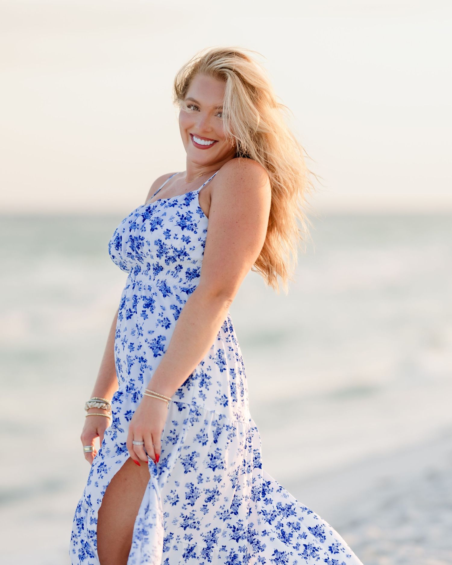 Smiling blonde woman twirls in a blue floral maxi dress on a sunny beach.