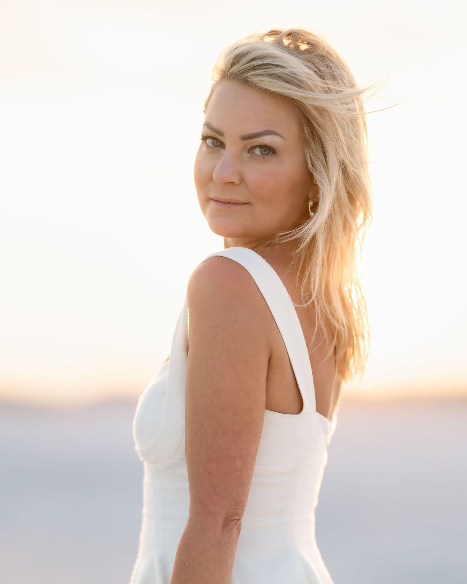 Blonde woman in white dress at sunset on a beach, golden hour light, looking over shoulder.