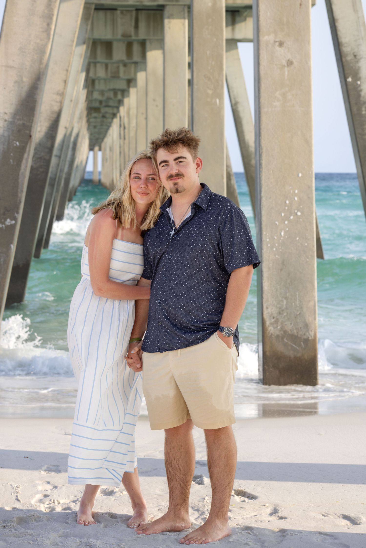 Couple standing barefoot under a wooden pier on a sandy beach, posing together by the ocean.