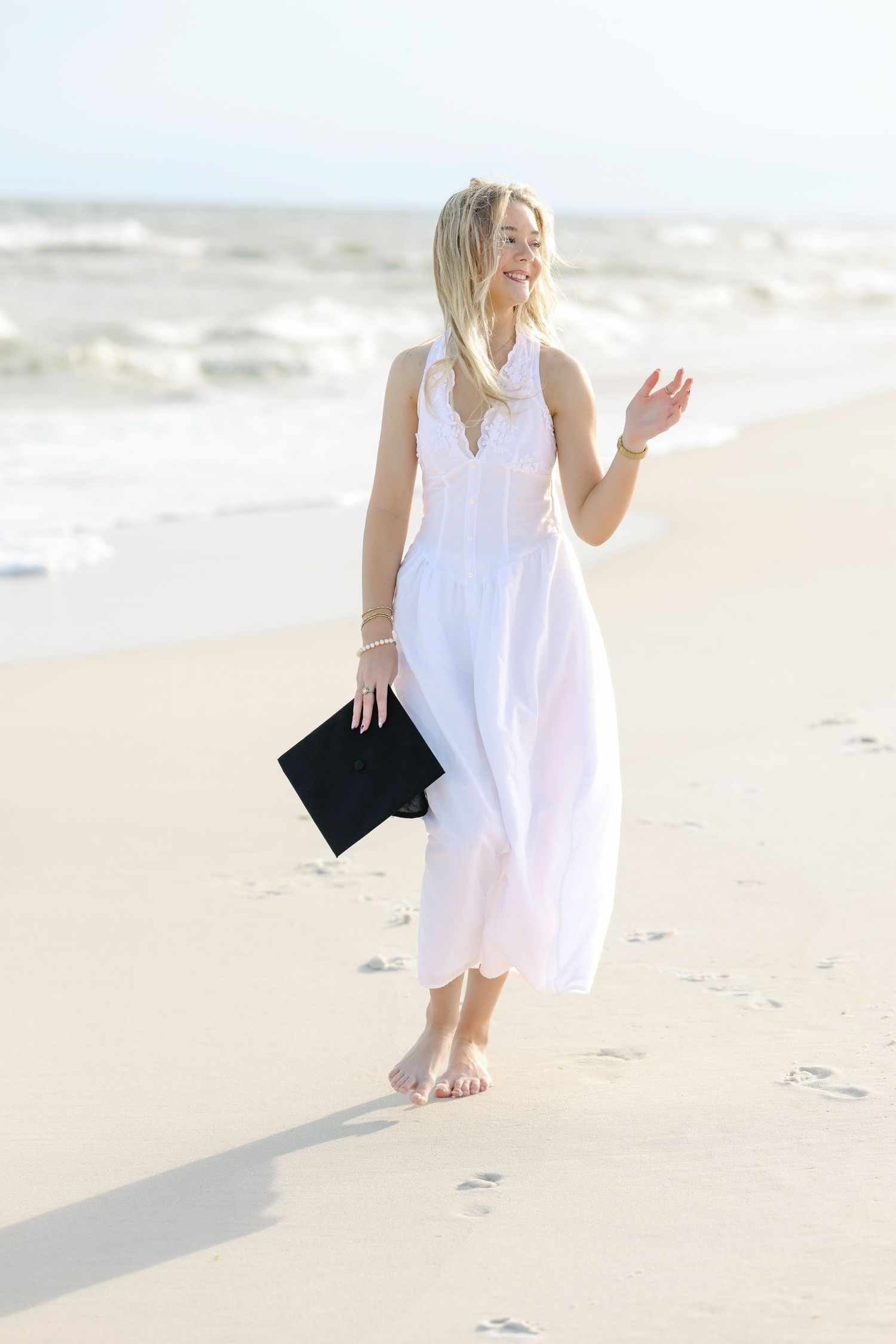 Joyful graduation photos on a sunny beach with waves and sand, featuring a white dress and black graduation gown.