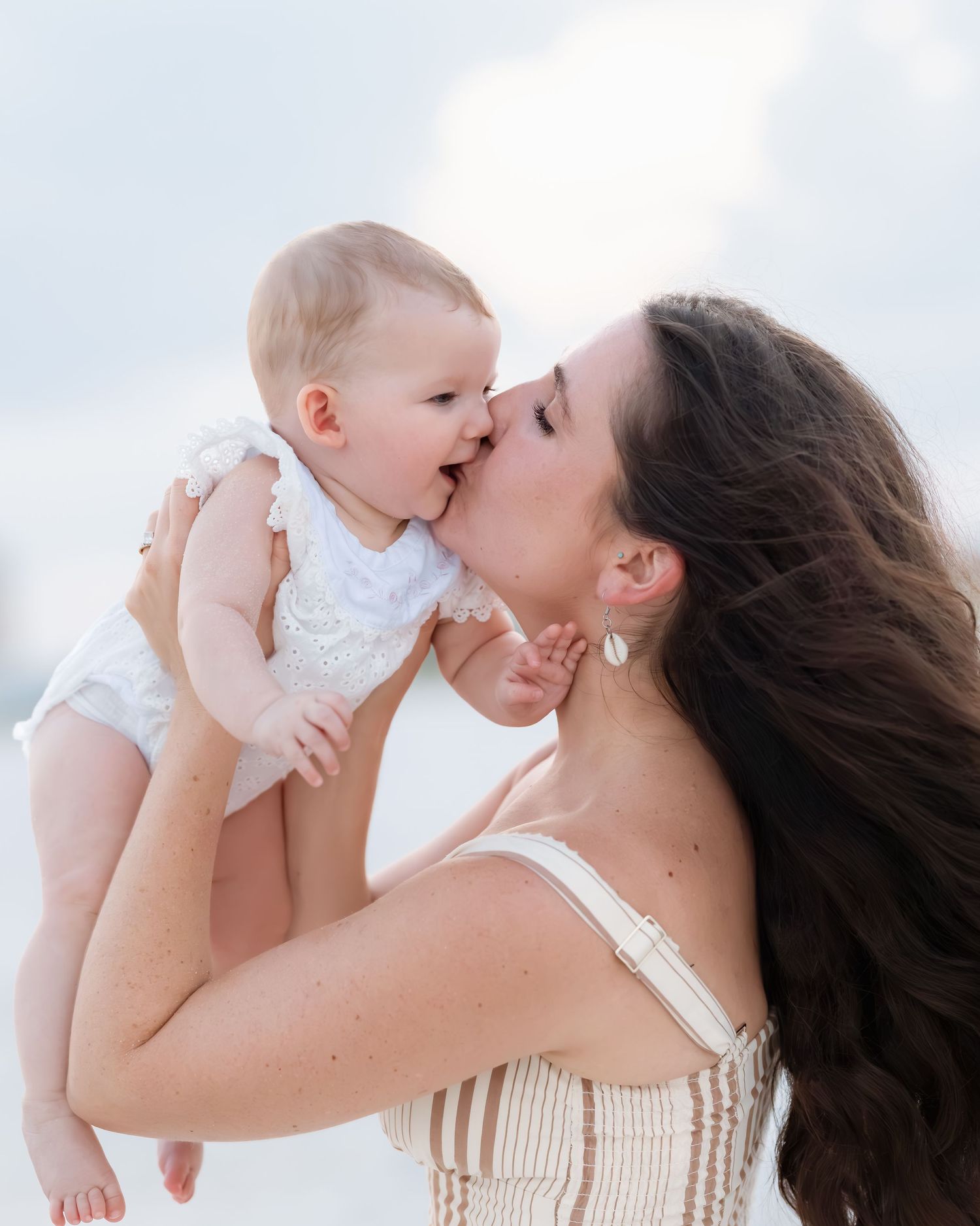 Mother kissing her smiling baby girl while holding her up against a bright sky at the beach.