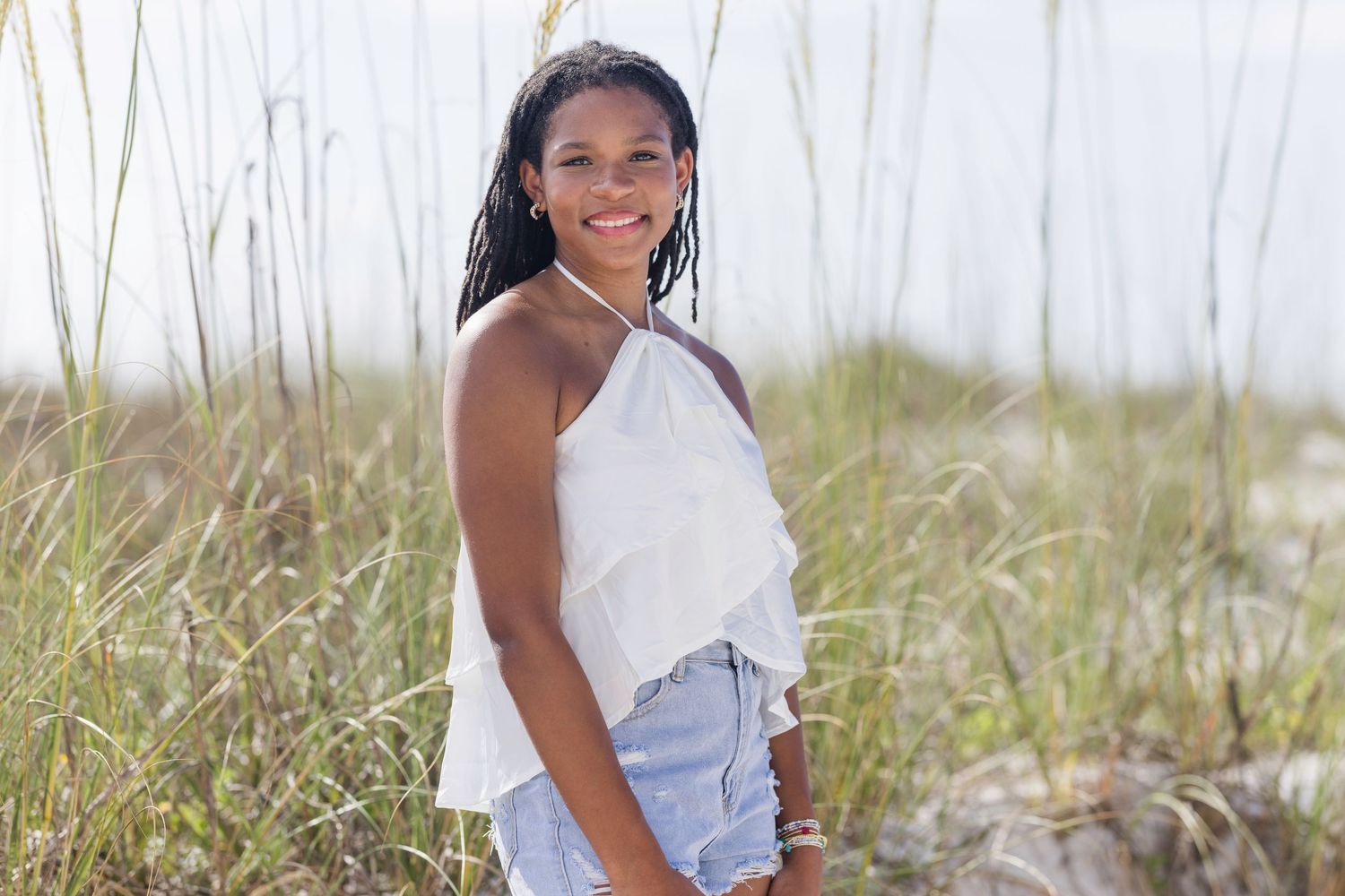 Young woman in white ruffled top and denim shorts smiling among beach dunes and sea grass.