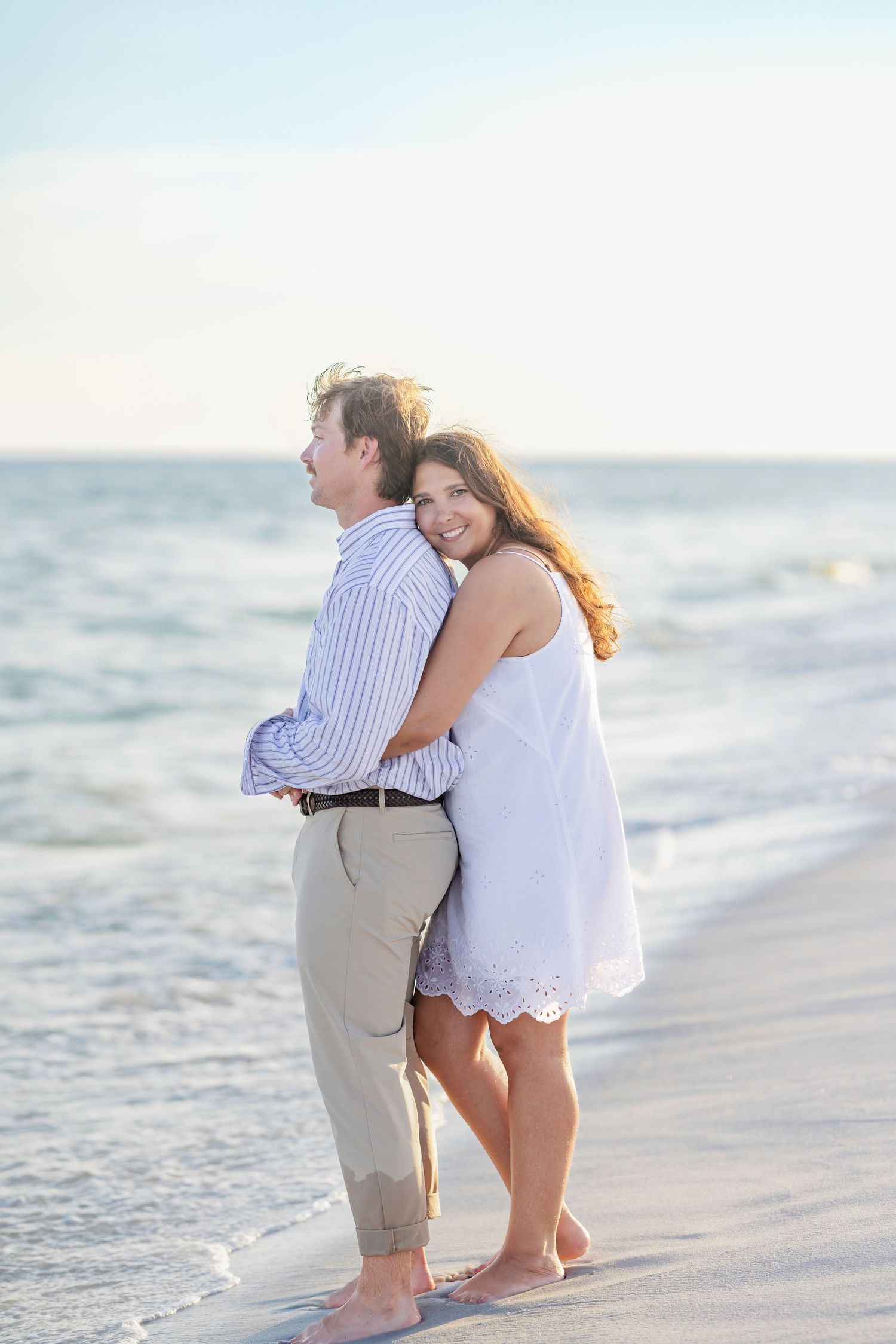 A couple embraces barefoot on a sunny beach, the woman in a white dress smiling back at the camera near the ocean.