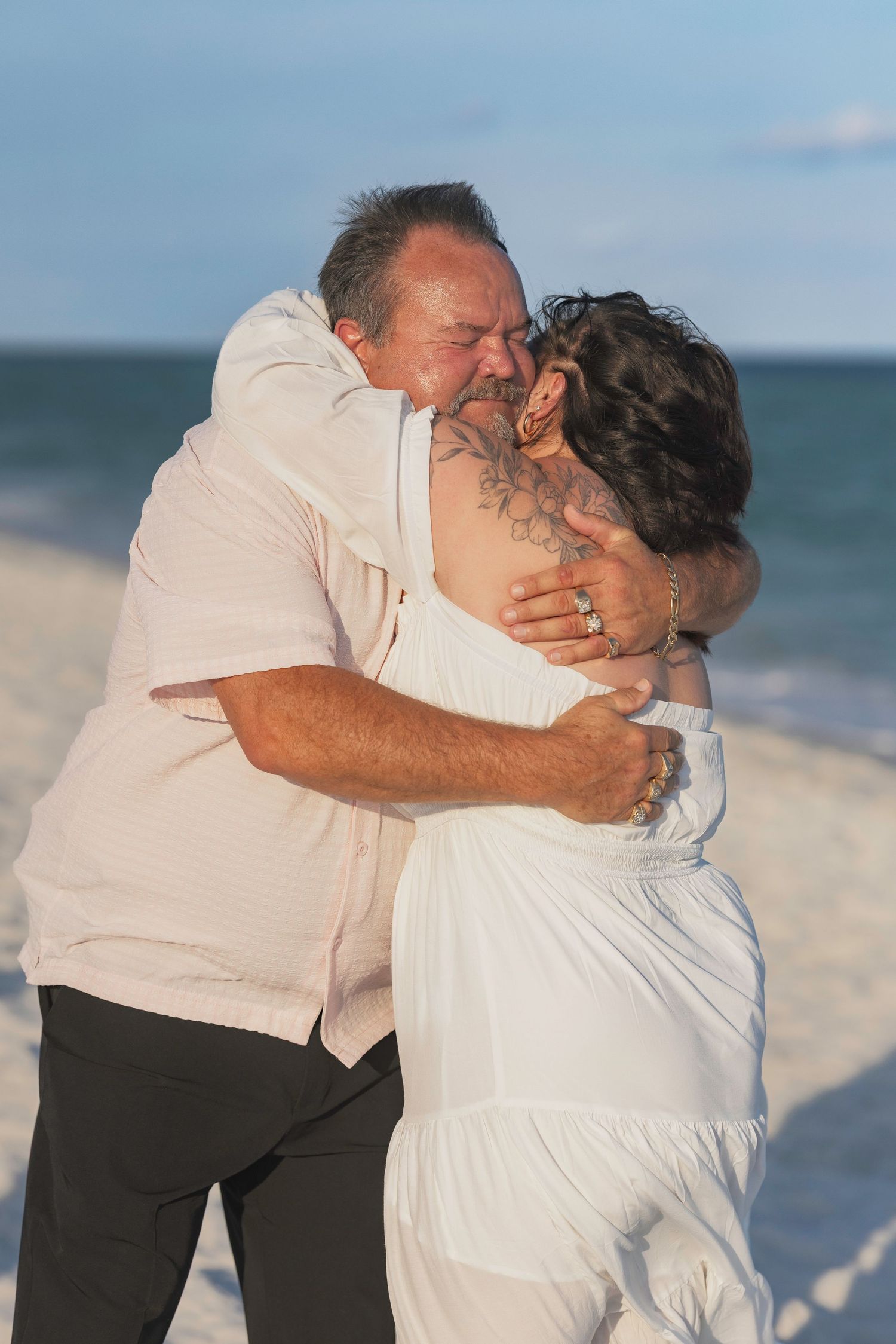 Couple embracing on a beach at sunset, wearing white outfits, sharing a romantic moment by the ocean.