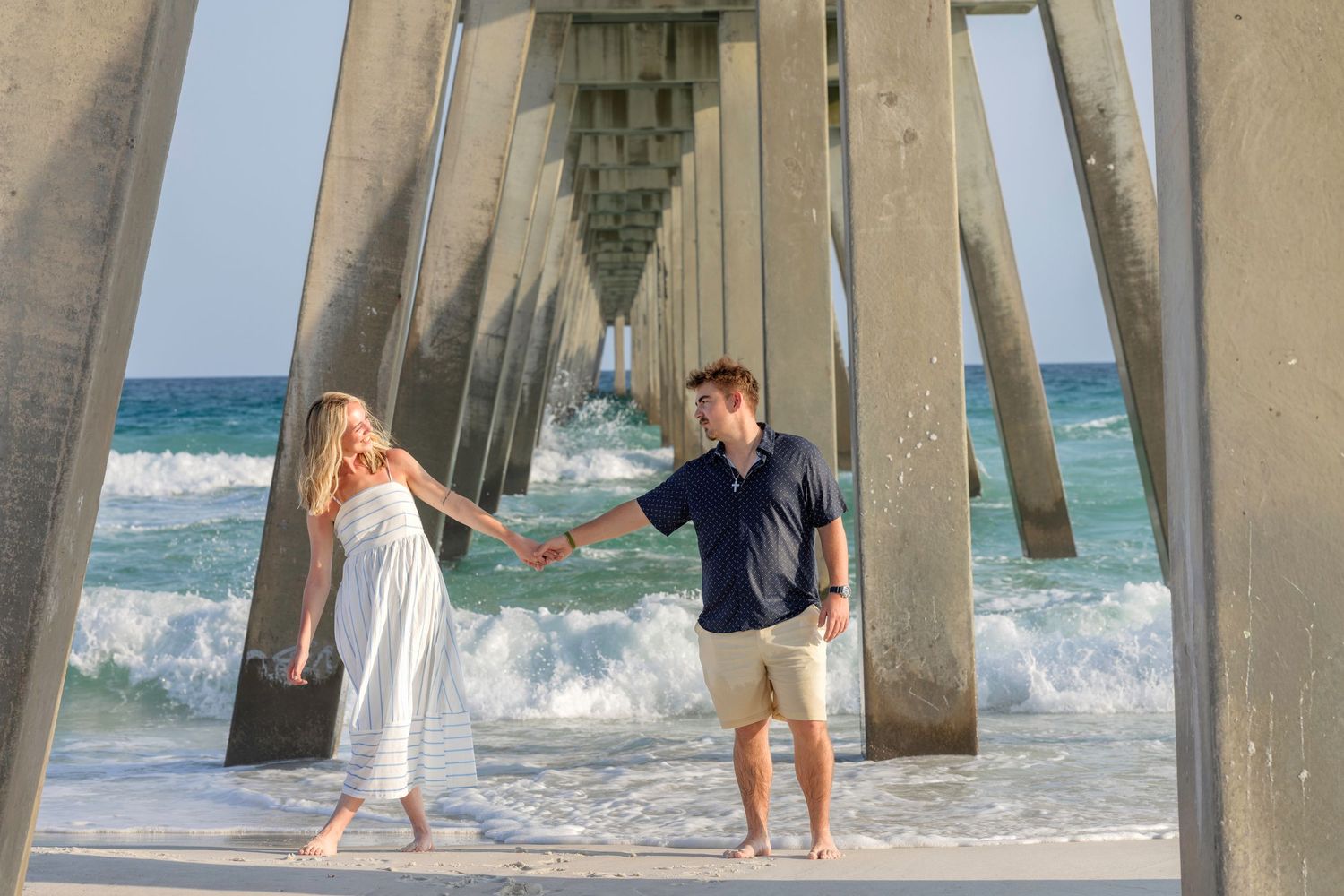Couple holding hands under a beach pier with ocean waves, white dress and navy shirt, romantic seaside photo.