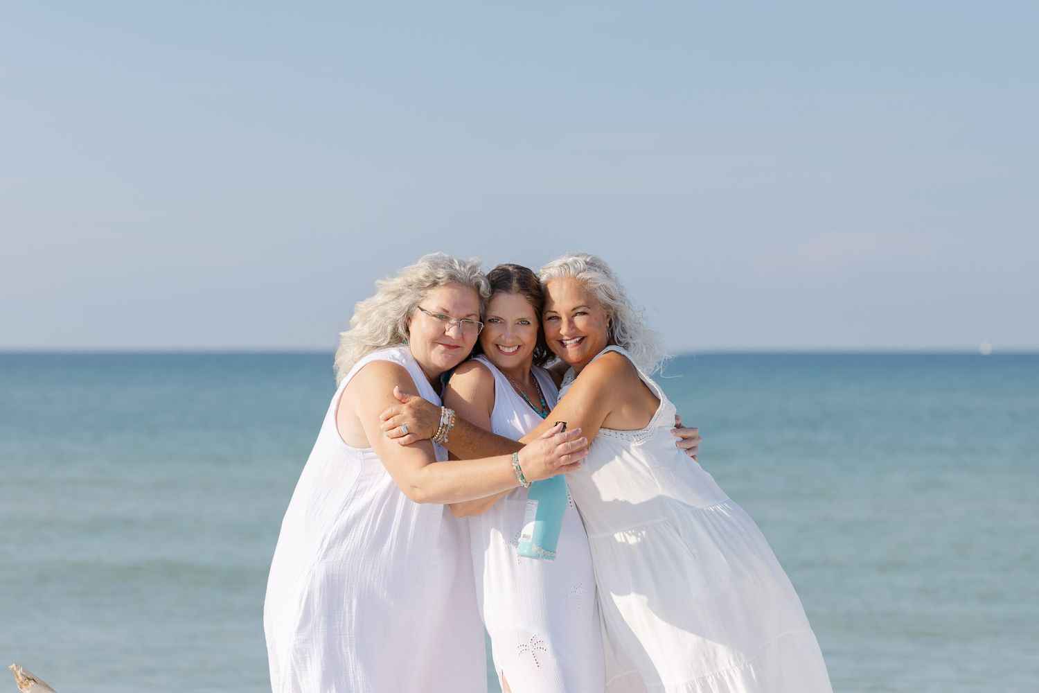 Family in white outfits embracing and smiling on a sunny beach with ocean waves in the background.