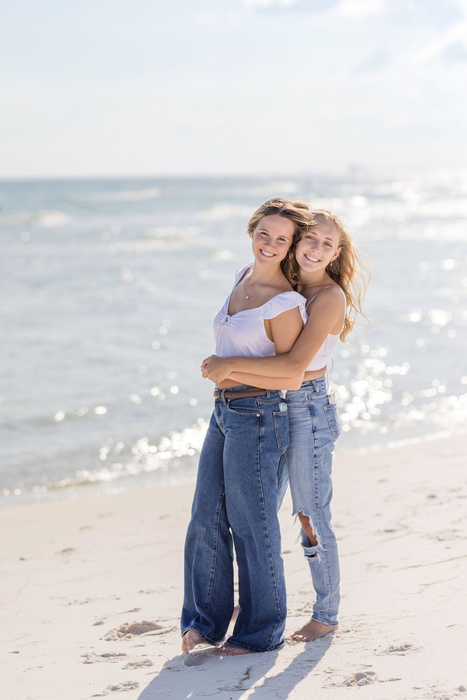 Two people embrace joyfully on a sunny beach with sparkling ocean waves in the background.