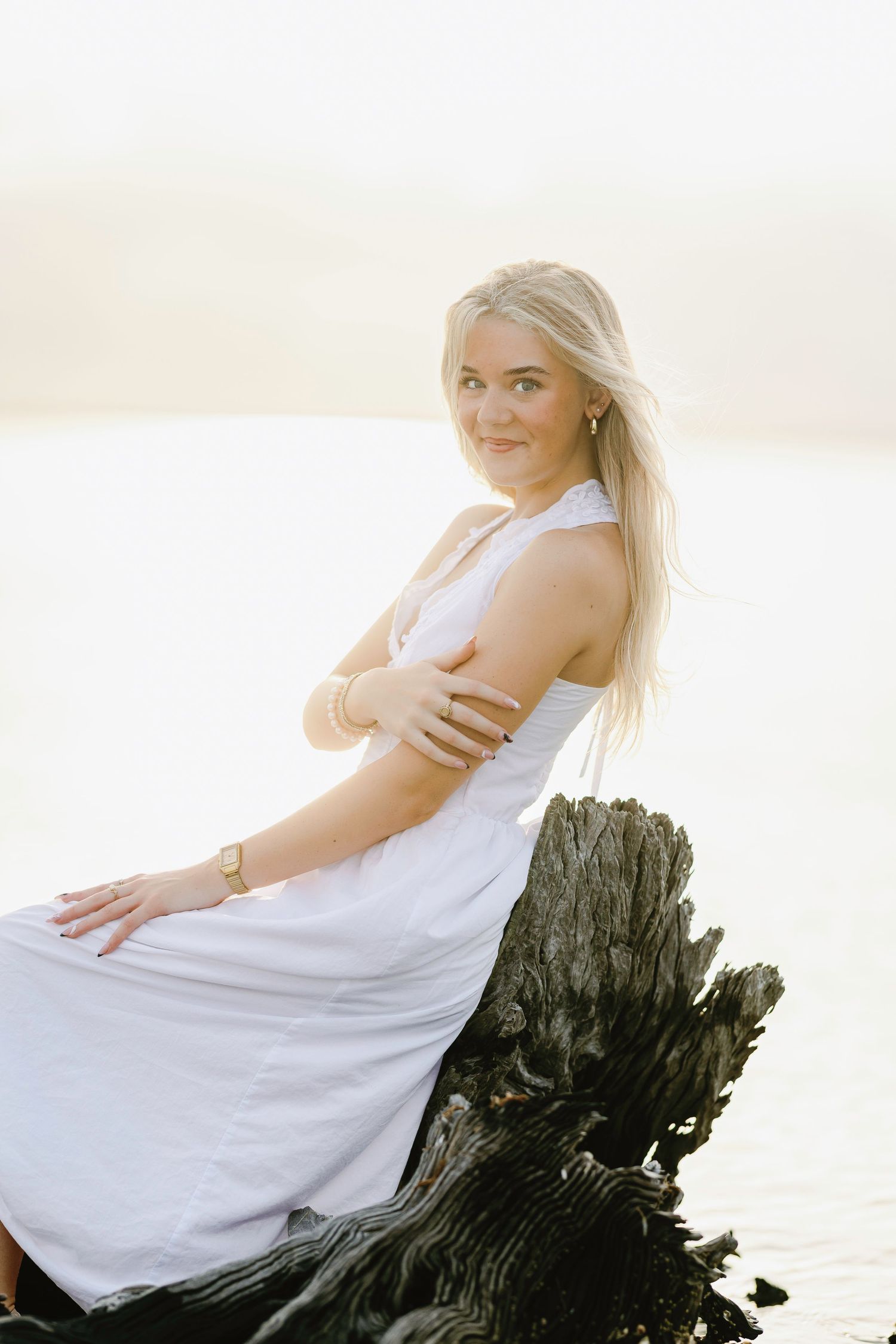 Woman in white dress poses elegantly on driftwood at beach during golden hour with soft lighting and ethereal atmosphere.