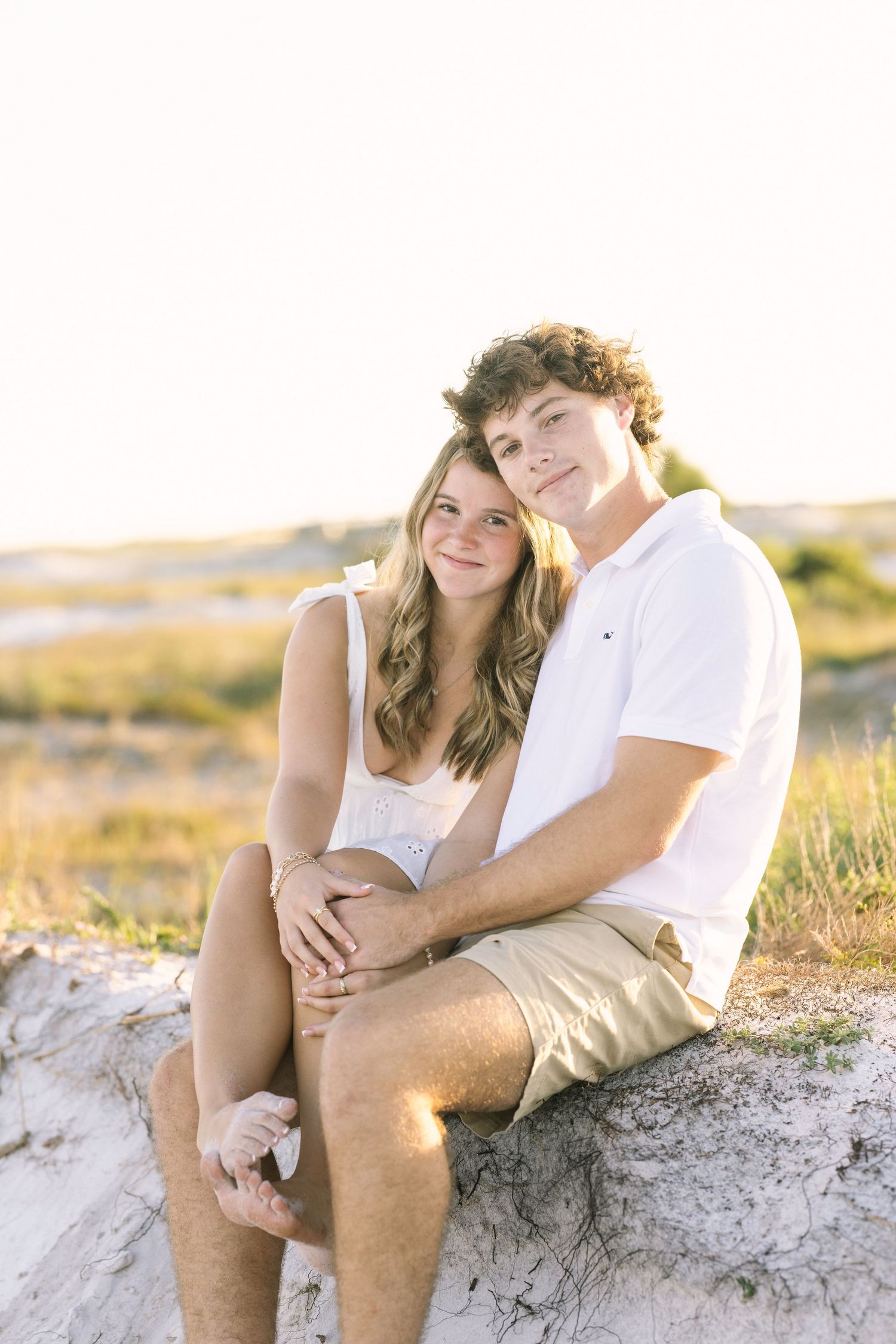 A couple sits together on rocks at the beach during golden hour as sunlight illuminates the natural landscape.