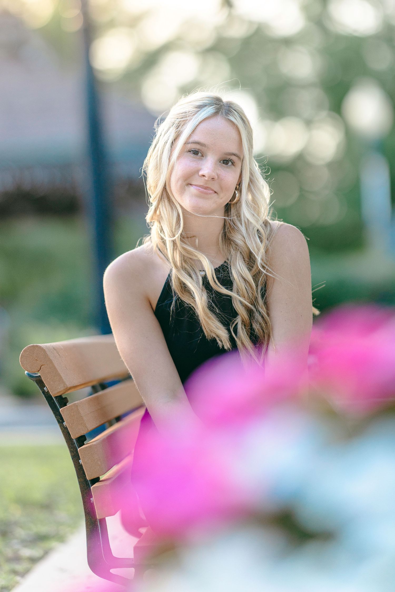Young blonde woman sitting on a wooden bench outdoors, smiling softly with pink flowers in the foreground.