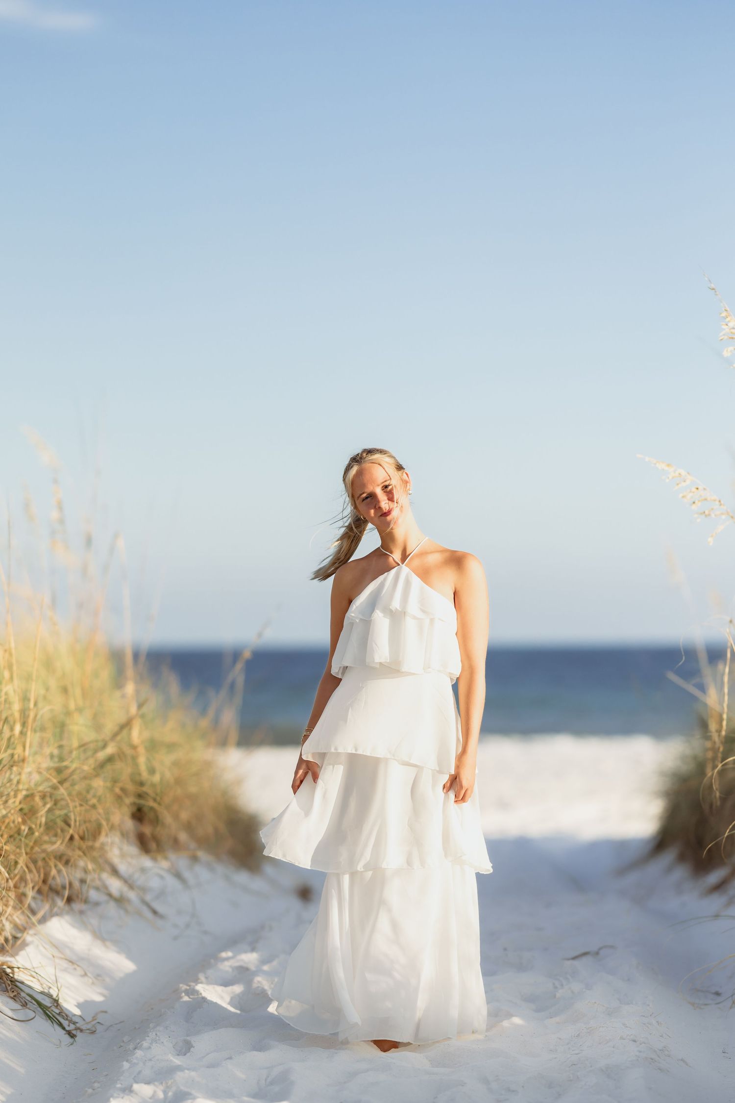 Smiling blonde woman in white tiered halter maxi dress standing on sandy beach path with sea oats and ocean backdrop.