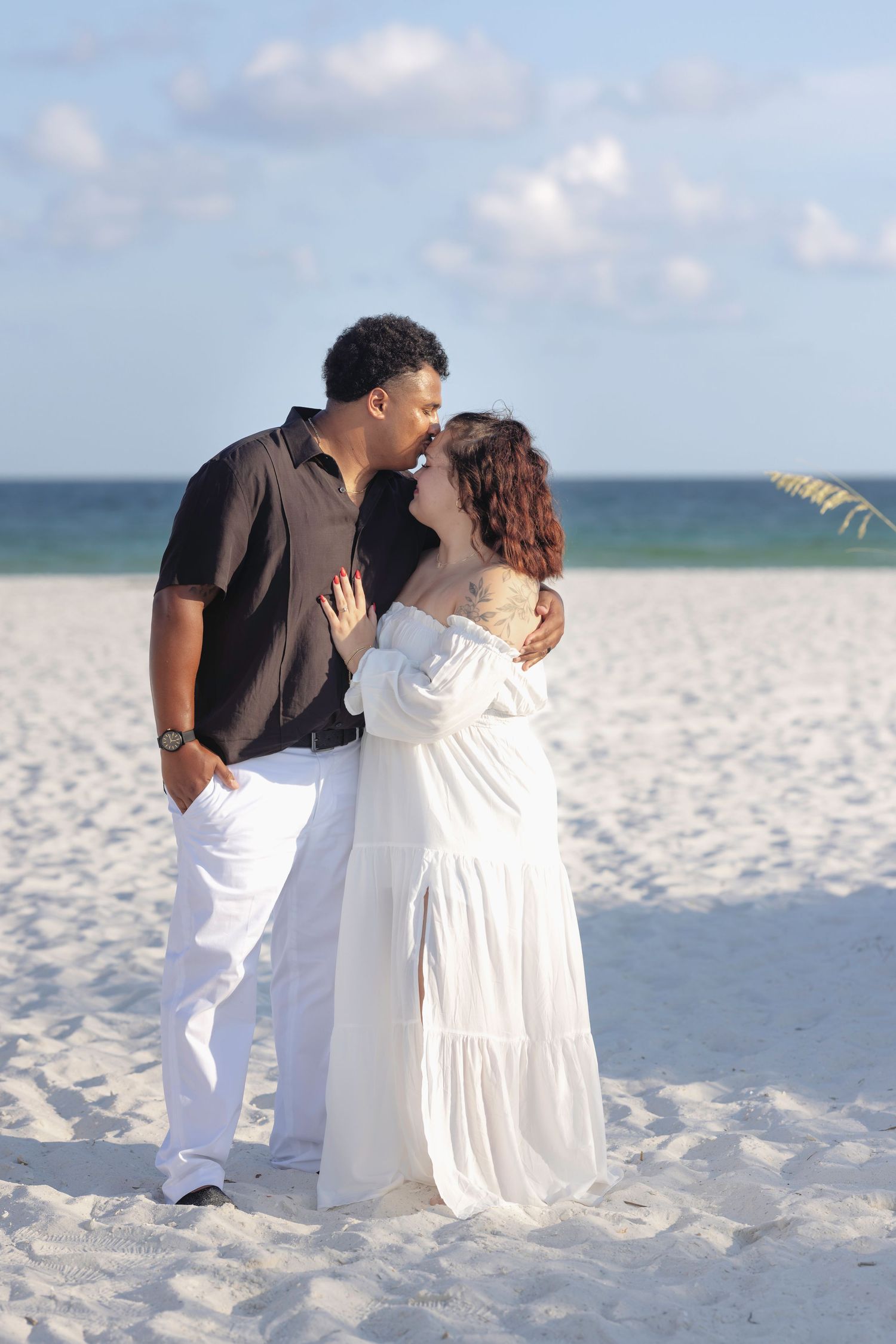 Couple sharing a romantic kiss on a white sandy beach, man in black shirt and woman in flowing white dress.