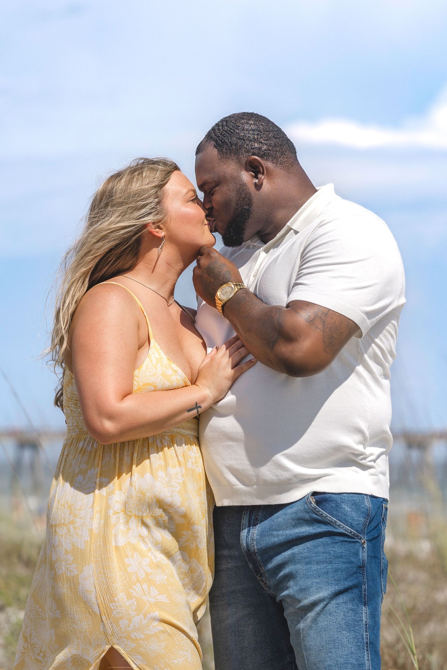 A couple shares a romantic kiss outdoors near a beach, woman in yellow dress, man in white shirt and jeans.