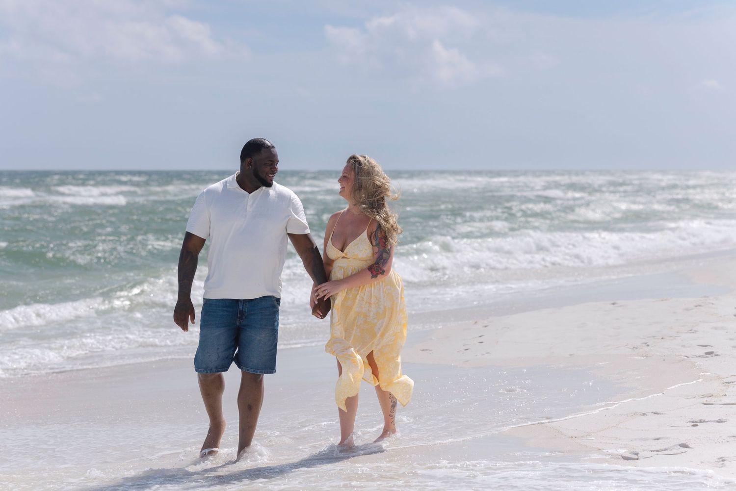 Couple walking hand-in-hand on a sunny beach, waves crashing behind them, man in white shirt and woman in yellow dress.
