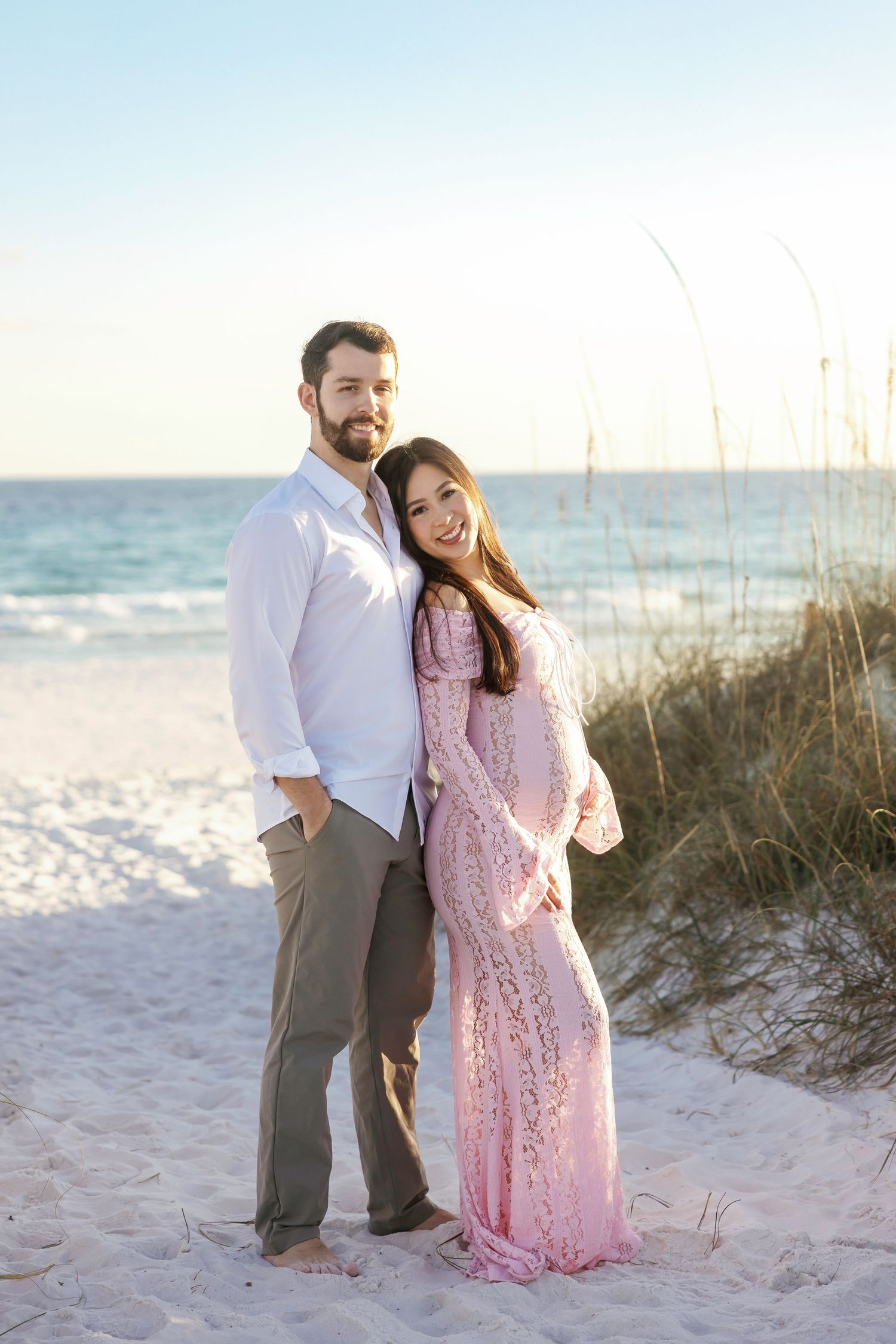 A couple shares romantic moments on a sandy beach with sea grass and ocean waves in the background.