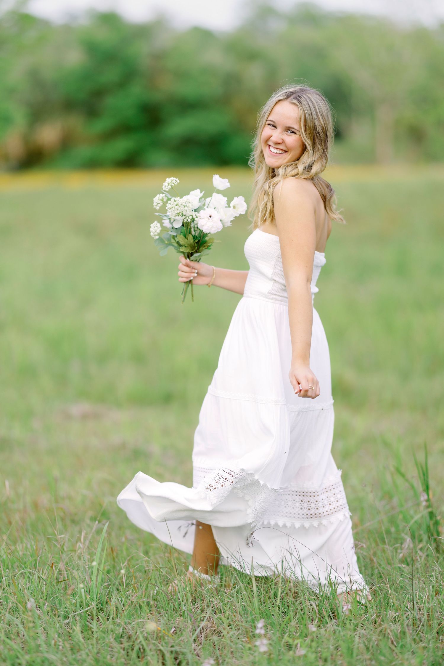 A bride in a flowing white dress holds a bouquet of white flowers while standing in a sunlit meadow.