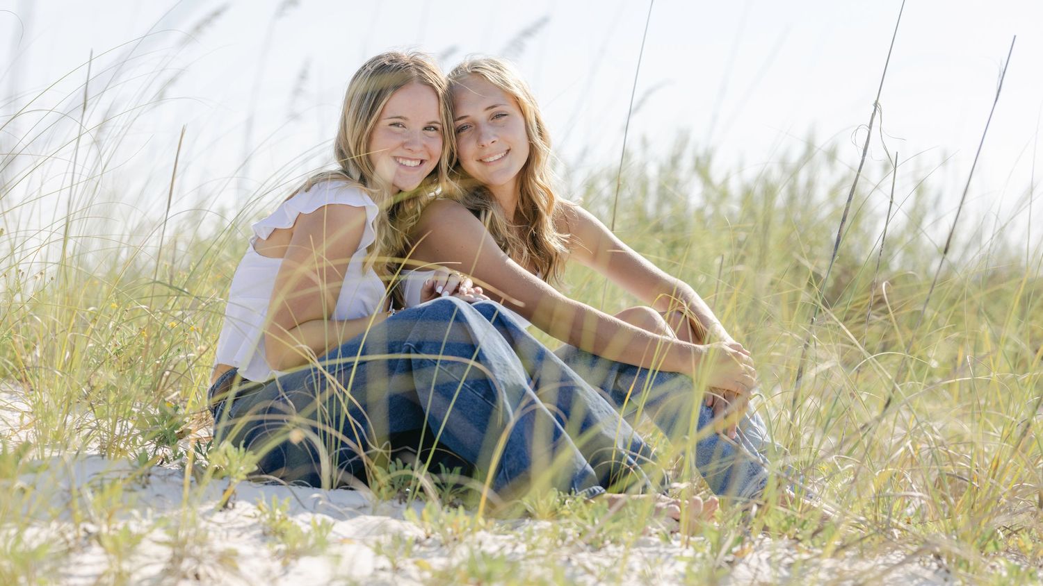 Two friends sit together smiling in a sunny field of tall grass on a bright summer day.