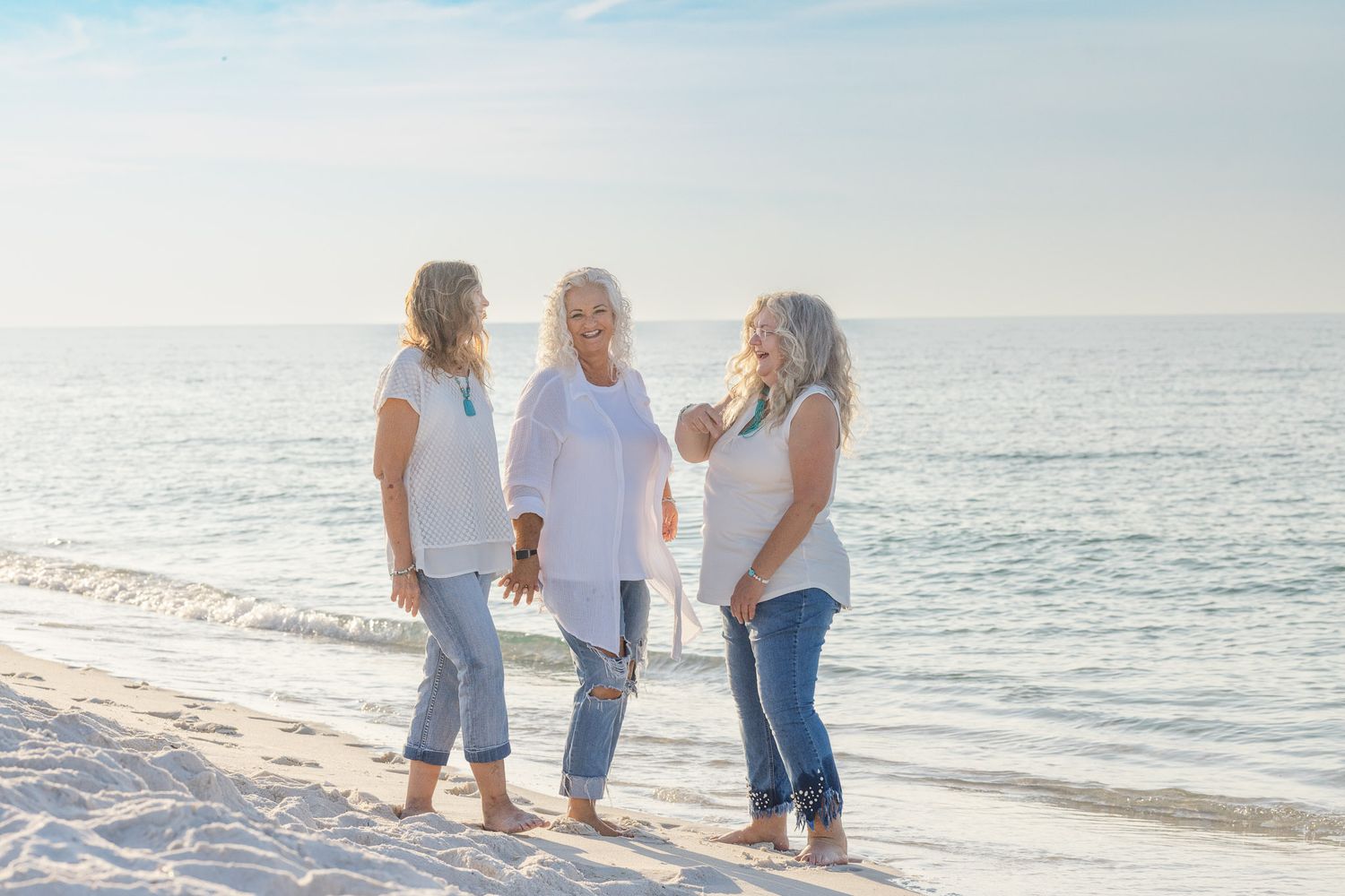 Three people stand together on a sunny beach wearing casual white and blue outfits with ocean waves in the background.