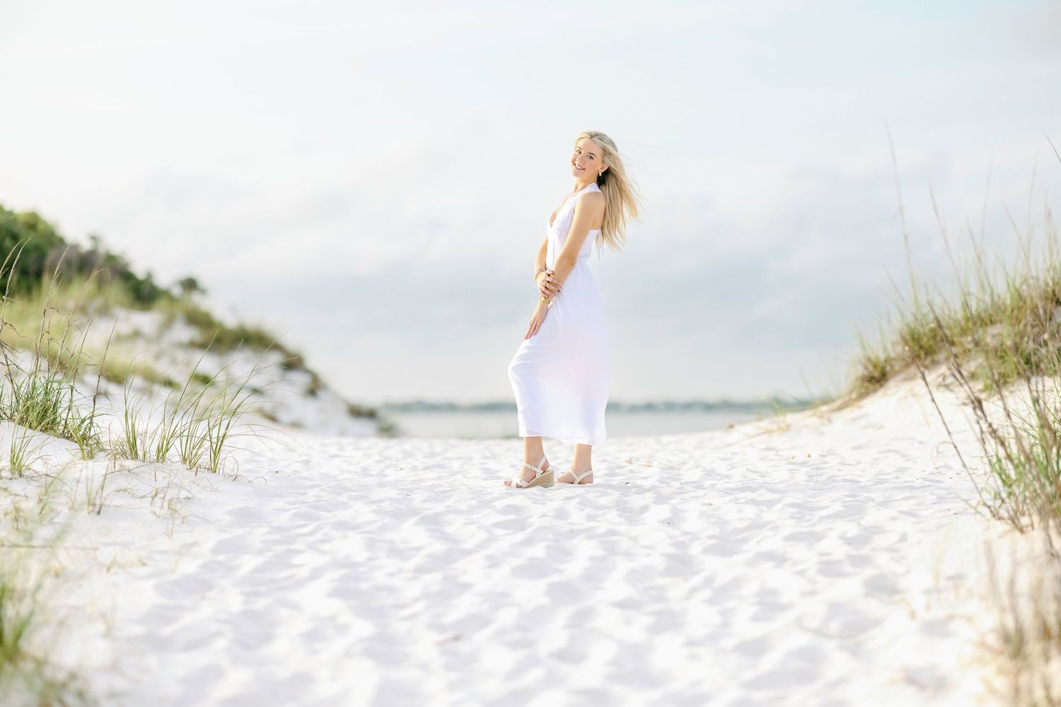 A figure in a flowing white dress stands on pristine white sand dunes by the ocean on a bright sunny day.