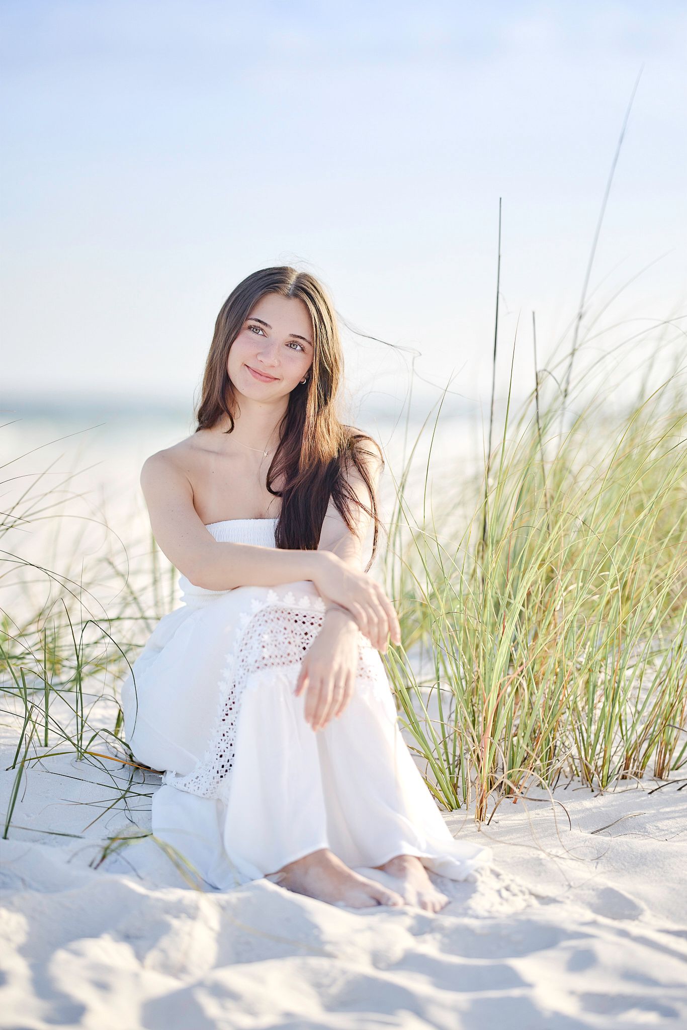 A dreamy beach portrait in a flowy white dress sitting among sand dunes and sea grass at sunset.