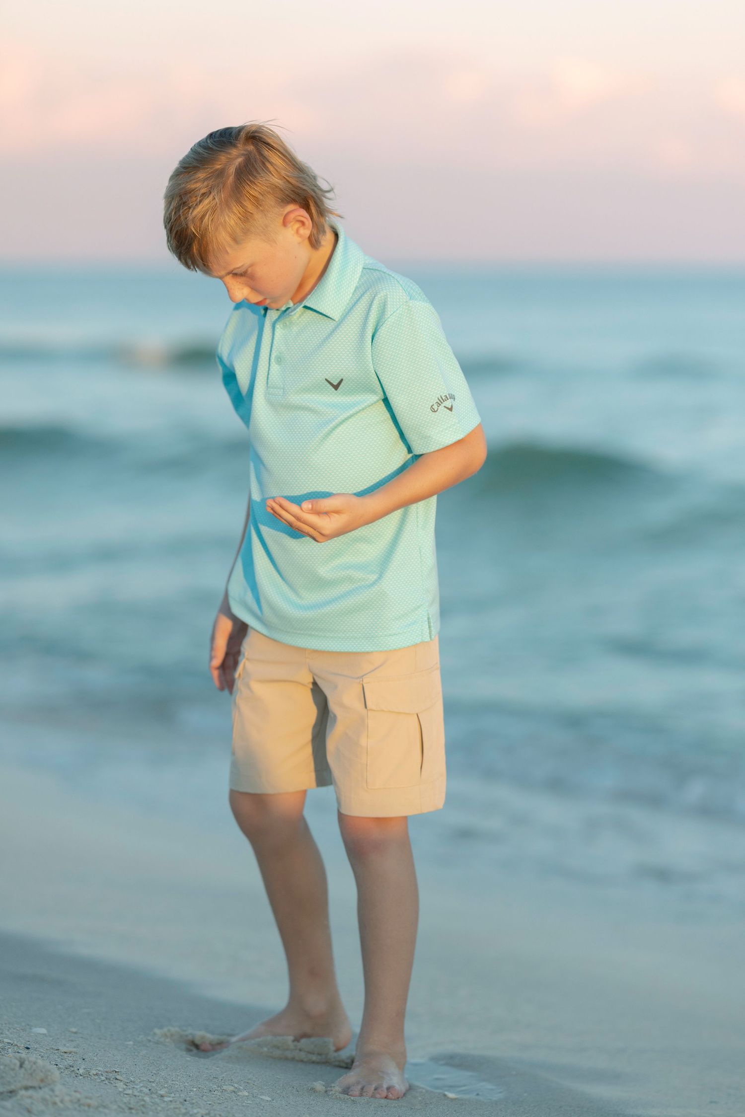 A child in a turquoise polo and khaki shorts looks down while standing barefoot on a beach at sunset.