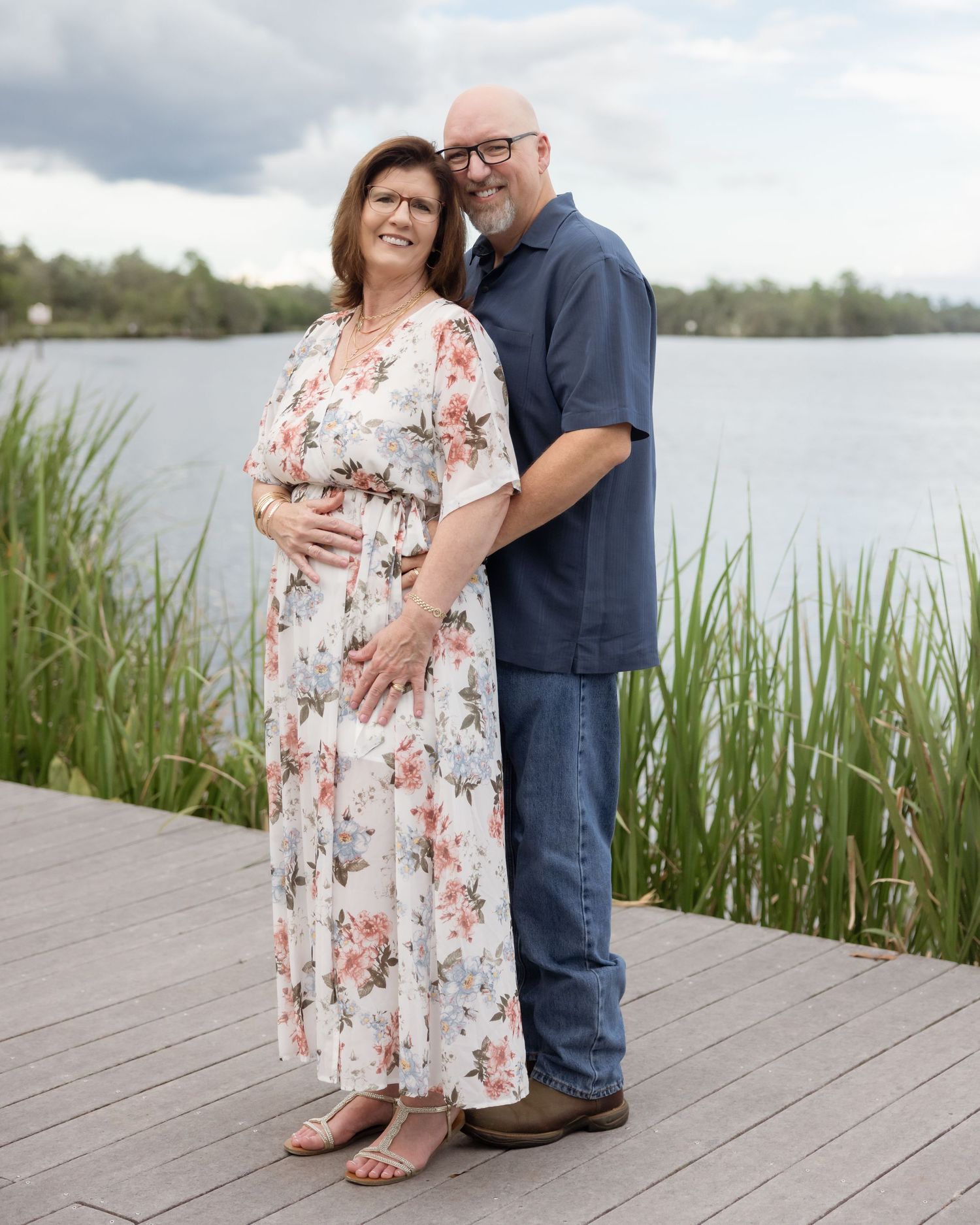 Expecting couple standing on a wooden dock by a lake, woman in floral dress cradling baby bump.