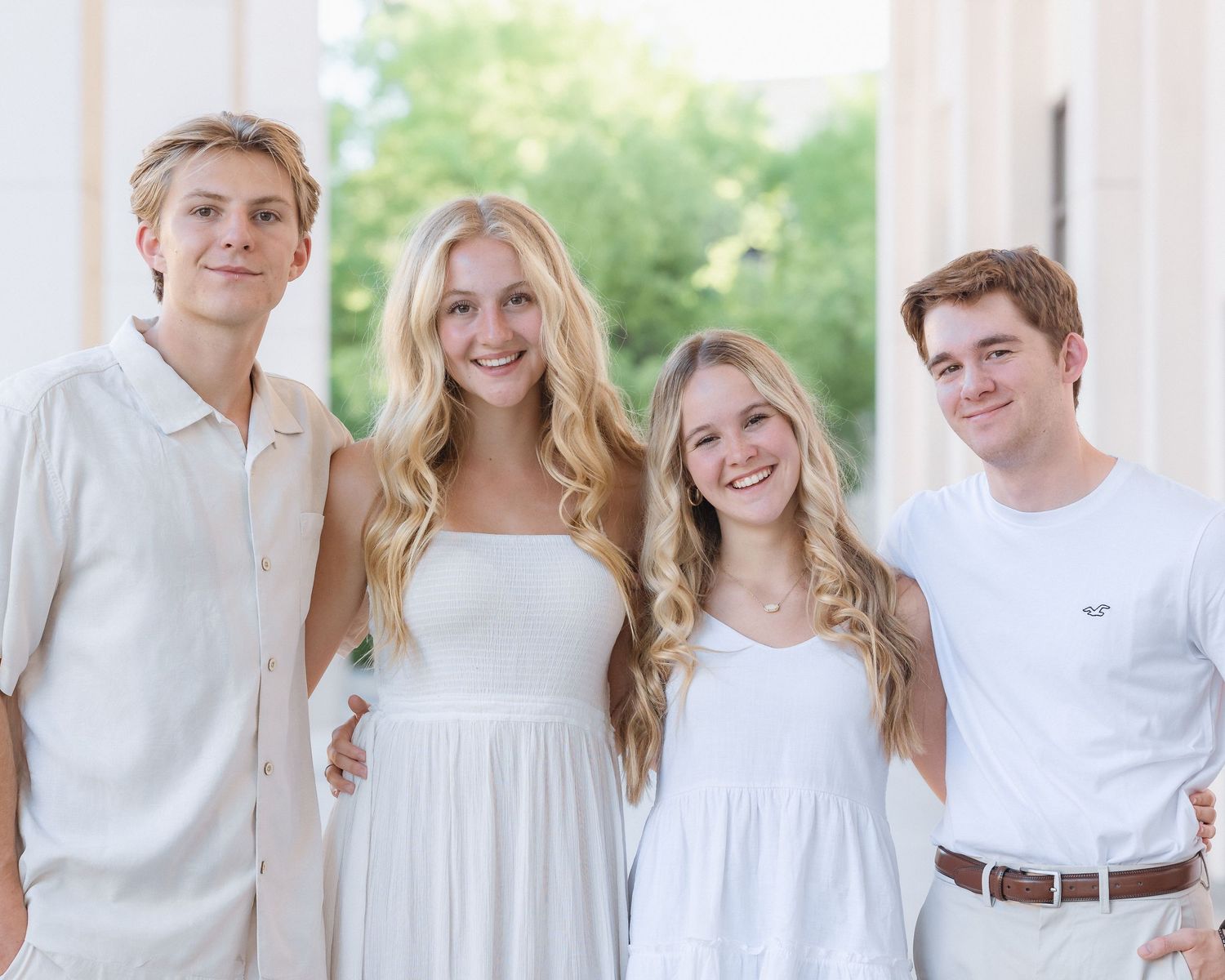 Four young adults smiling together in white outfits outdoors.