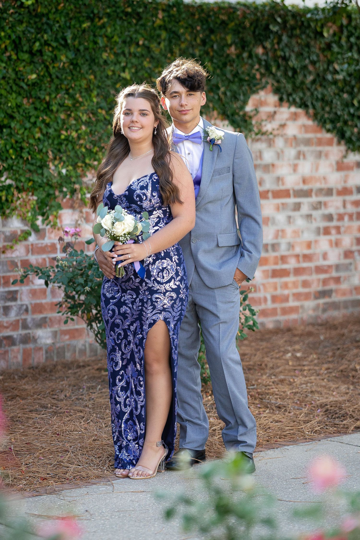 A teenage couple poses in prom attire, she in a blue gown with bouquet, he in a gray suit with boutonniere.