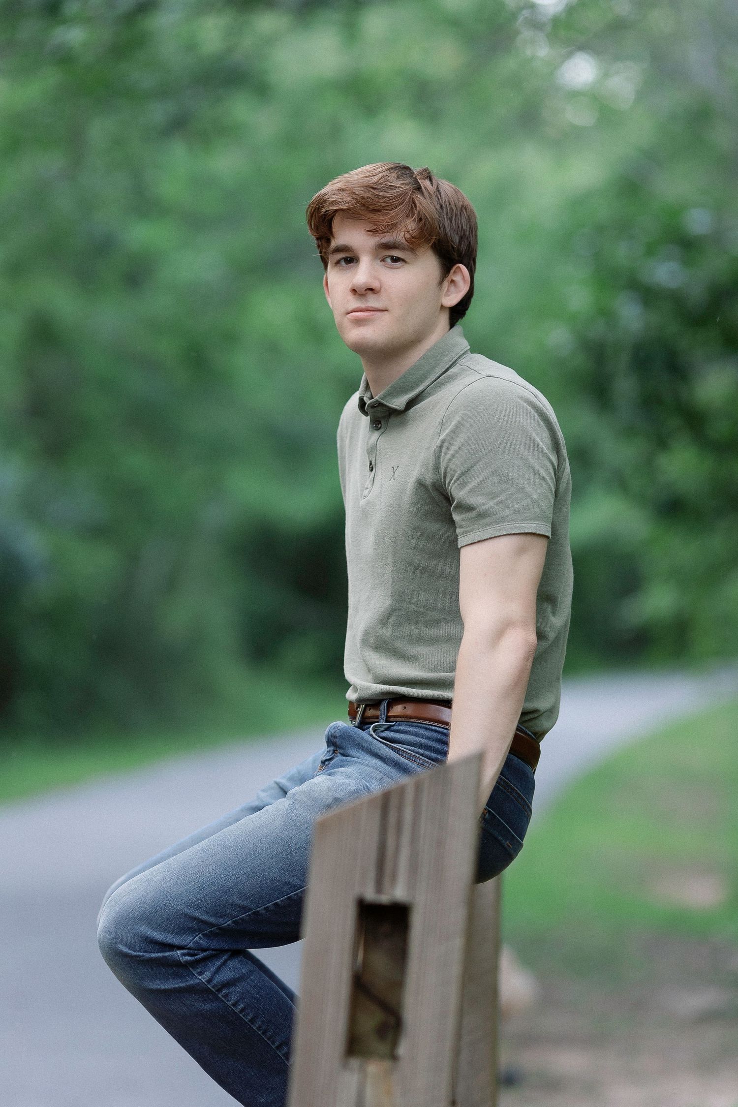 Young man with auburn hair sits on wooden fence in jeans and olive polo shirt along a tree-lined path.