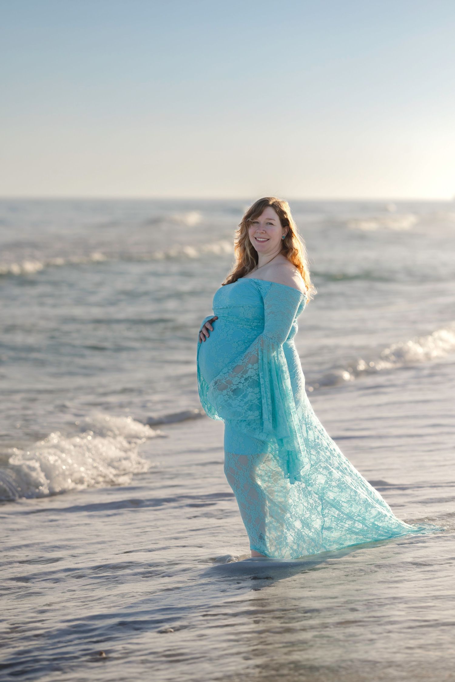 Pregnant woman in teal lace gown standing in ocean waves on a sunny beach, smiling and cradling her baby bump.