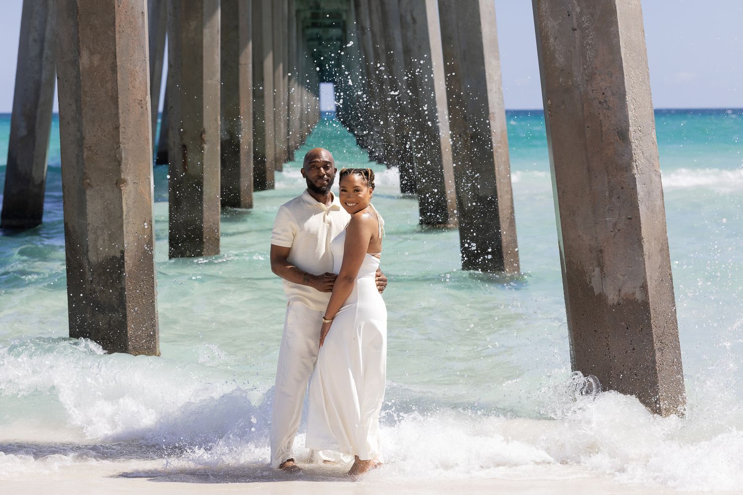 Couple in white outfits embracing under a beach pier with ocean waves splashing around them.