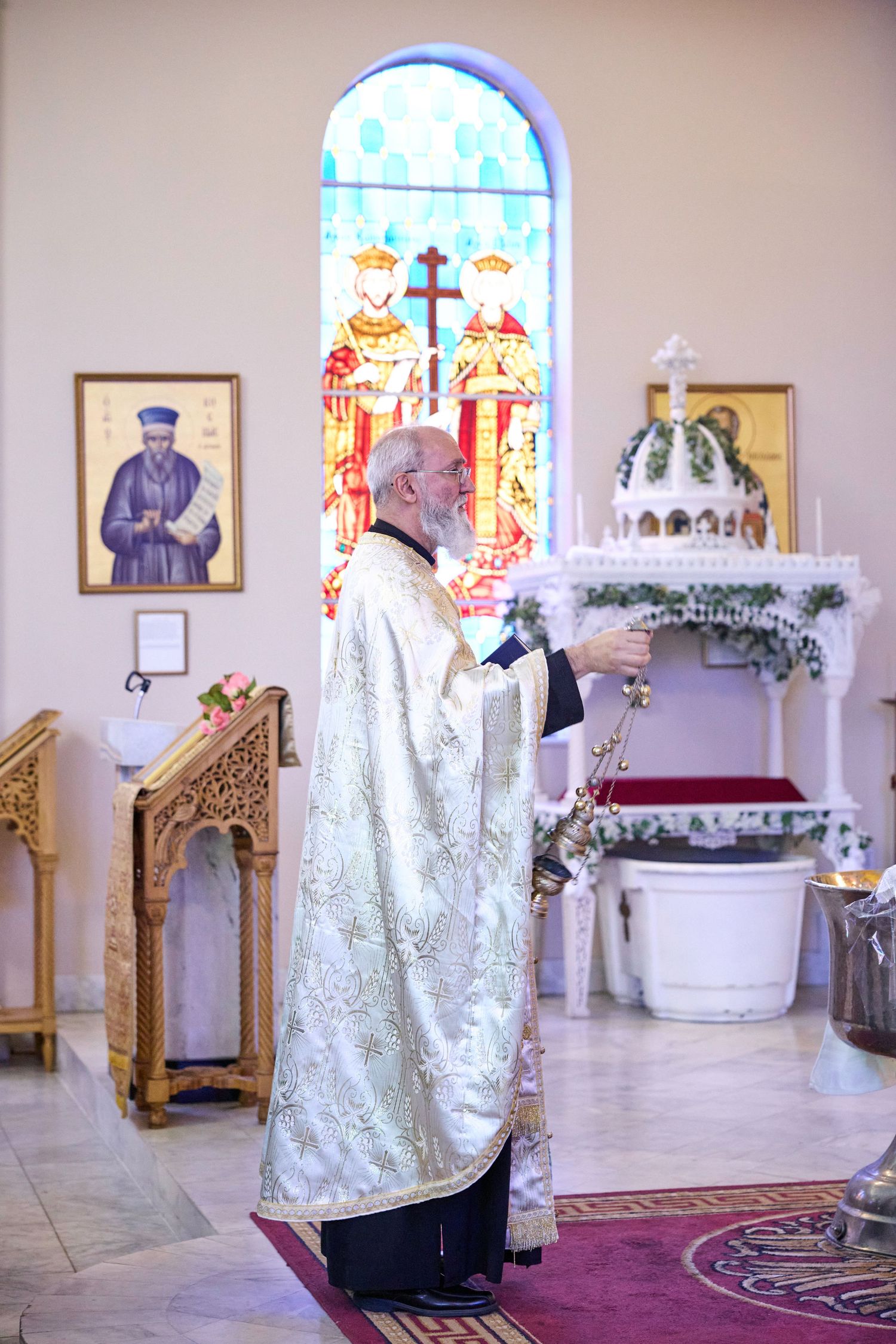 Orthodox priest in white and gold vestments performs religious ceremony at ornate church altar with stained glass windows.