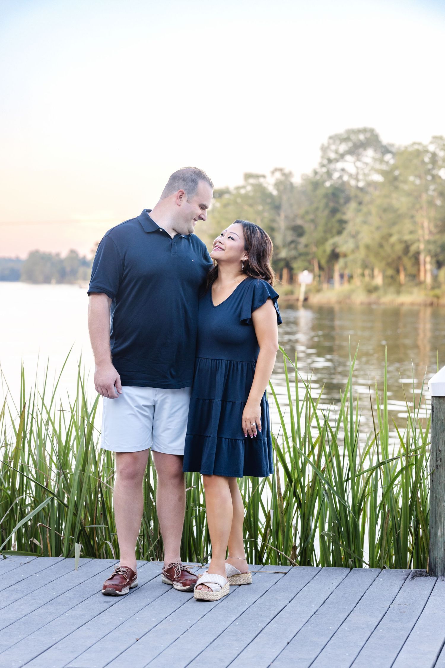 A couple shares a joyful moment together on a wooden dock by a serene lake surrounded by tall grass at sunset.