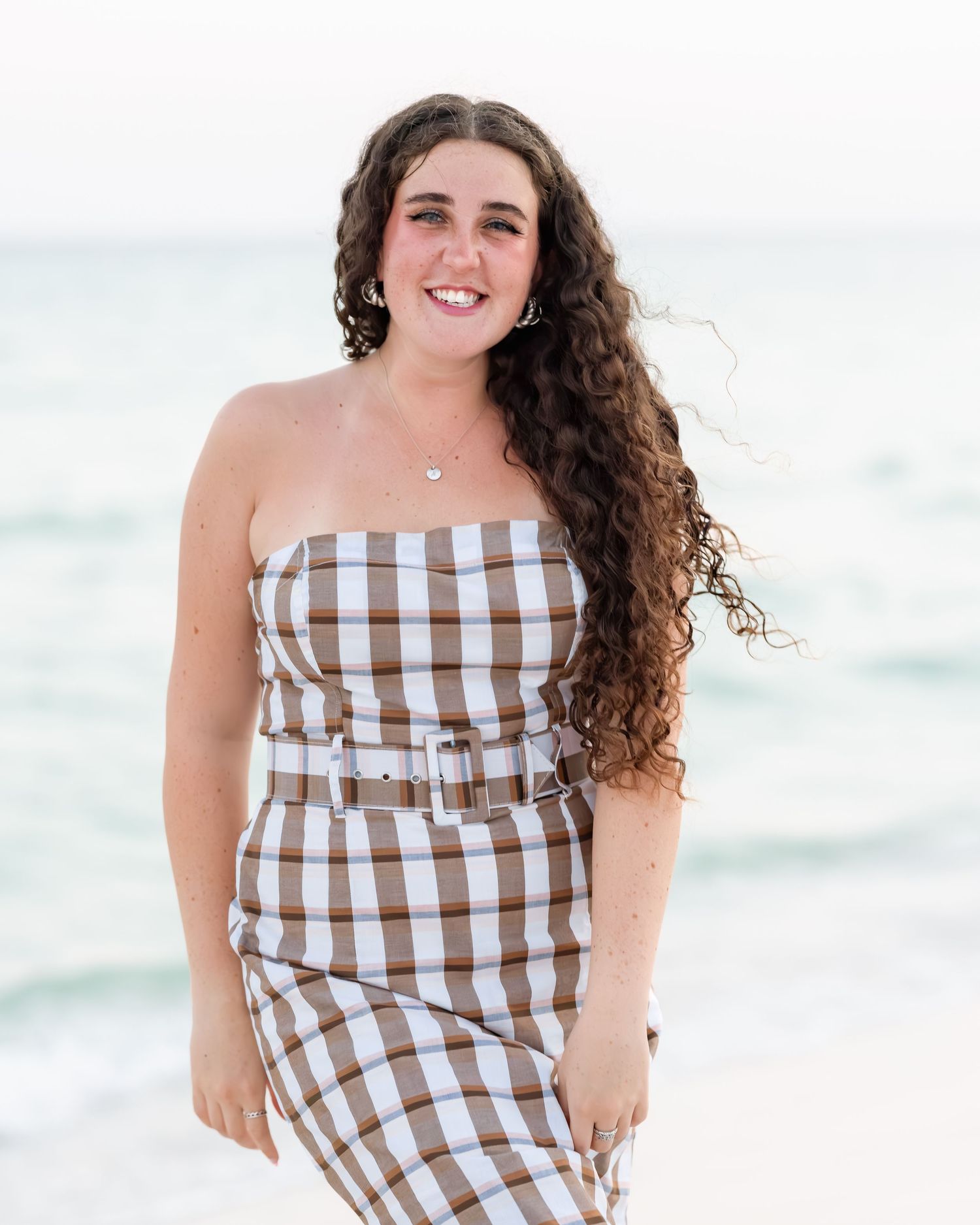 Young woman with long curly hair smiles confidently in brown plaid strapless dress on beach.
