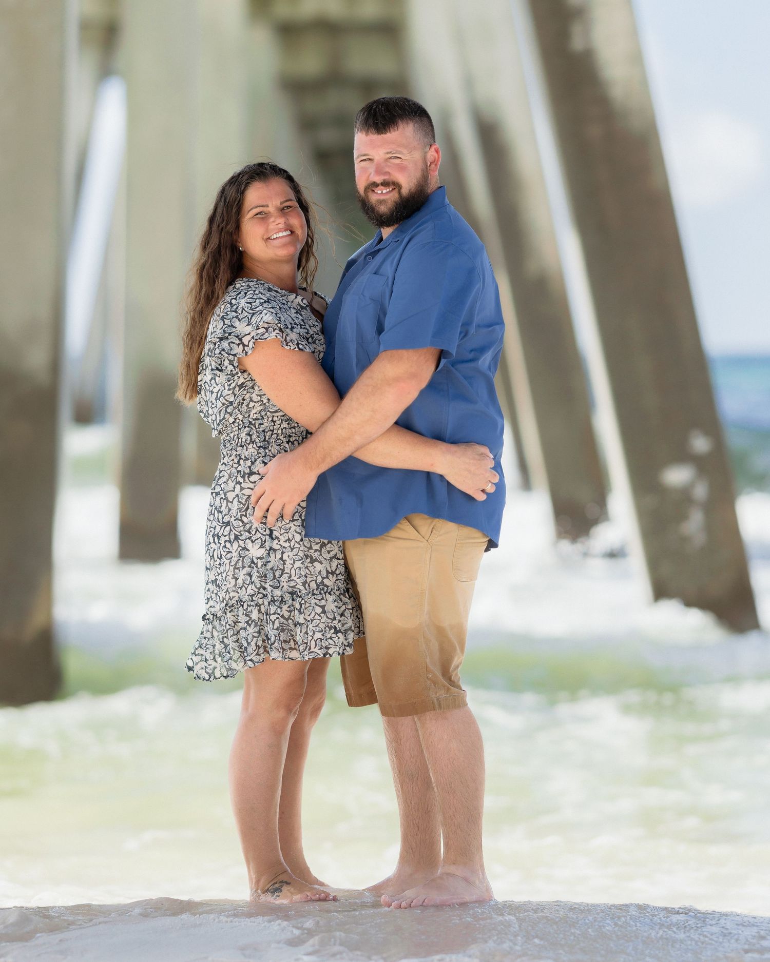 Couple embracing and smiling on a beach near a pier, woman in floral dress and man in blue shirt and khaki shorts.