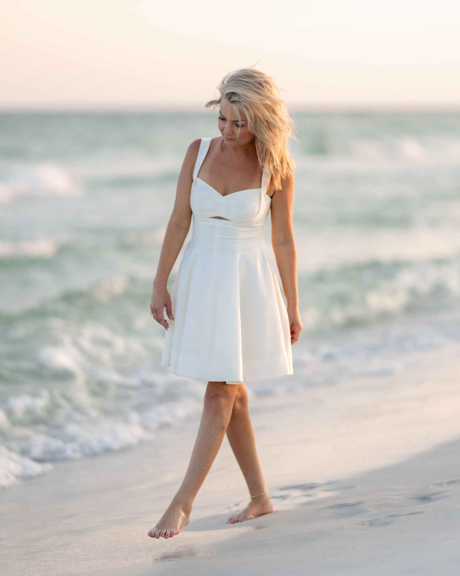 Blonde woman in white sundress walking barefoot on sandy beach with ocean waves in background.