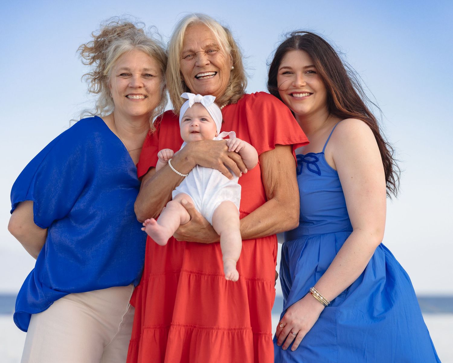 Three women in red and blue outfits smile together at the beach, with the middle woman holding a baby in white.
