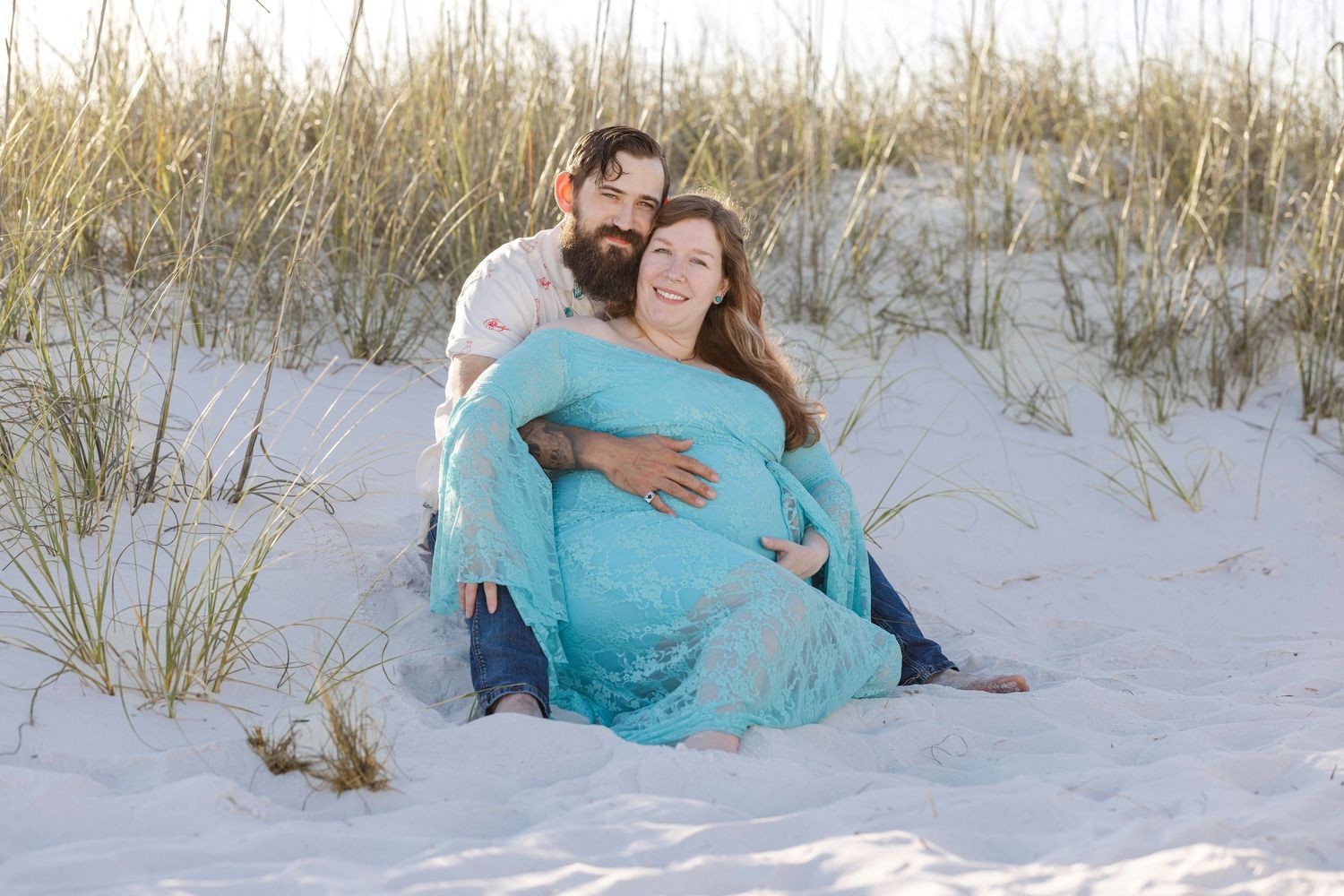 Pregnant couple sitting on white sand beach dunes, woman in teal maternity gown, man embracing her from behind.