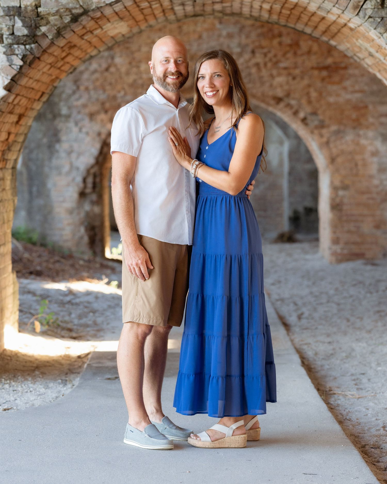 Smiling couple poses under brick arches, man in white polo and khaki shorts, woman in blue maxi dress and wedge sandals.