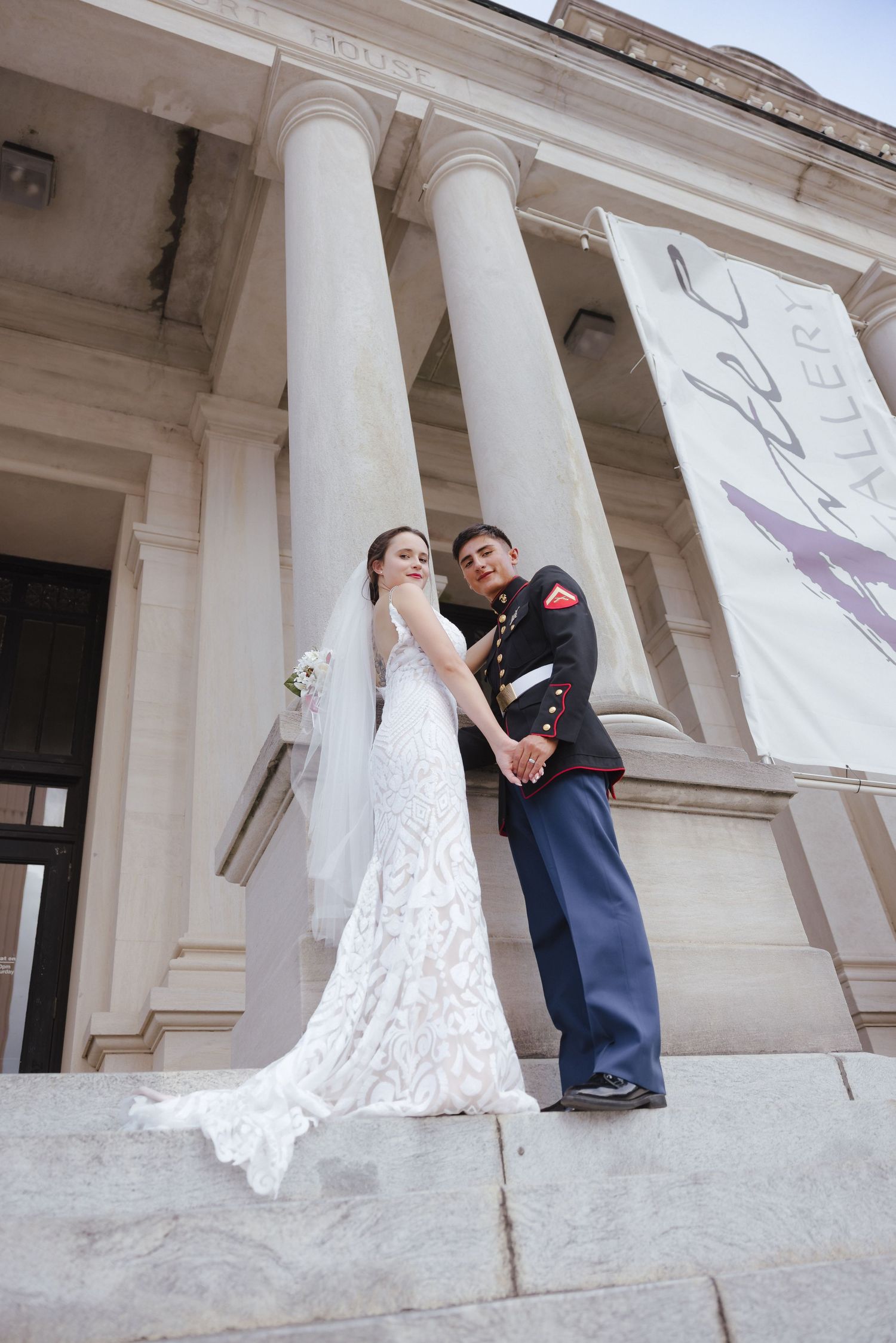 Bride in white lace gown and groom in Marine dress uniform hold hands on steps of elegant columned building.