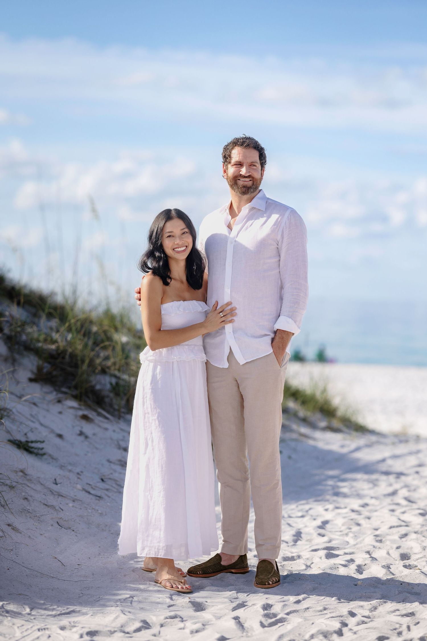 A smiling couple in white and beige outfits poses on a sandy beach with blue sky and dunes behind them.