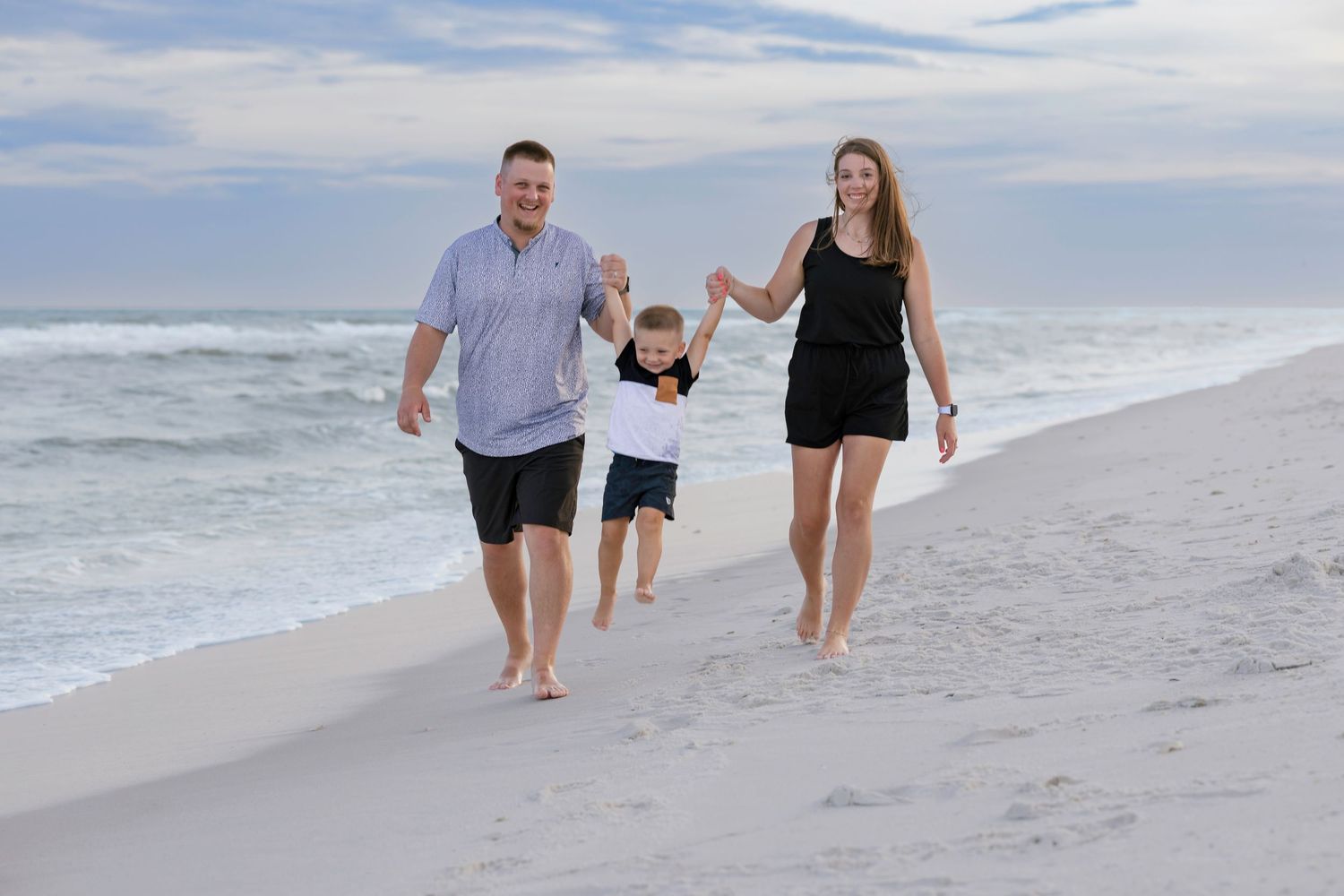 Family of three walking on white sand beach near ocean waves, parents holding child's hands, cloudy sky backdrop.