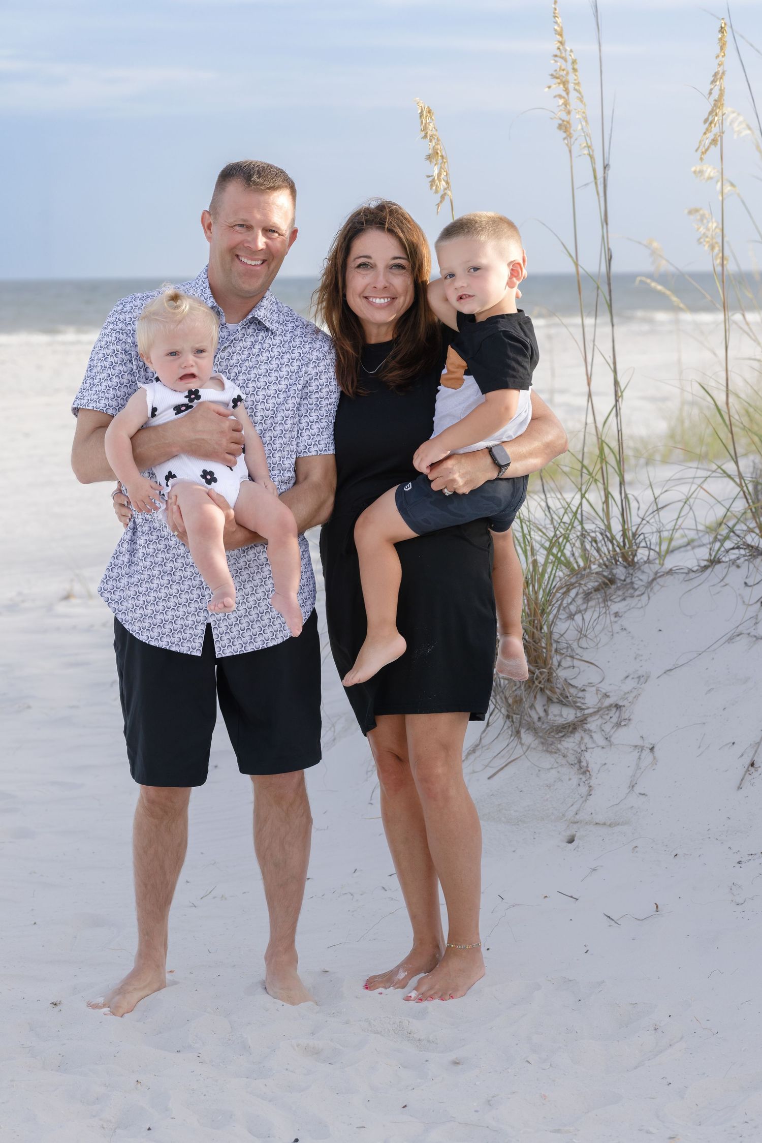 Happy family of four posing on a white sand beach, parents holding two young children near sea oats dunes.