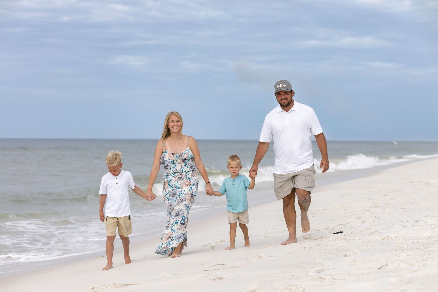 Family of four walking hand-in-hand on a sunny beach, with waves and blue sky in the background.