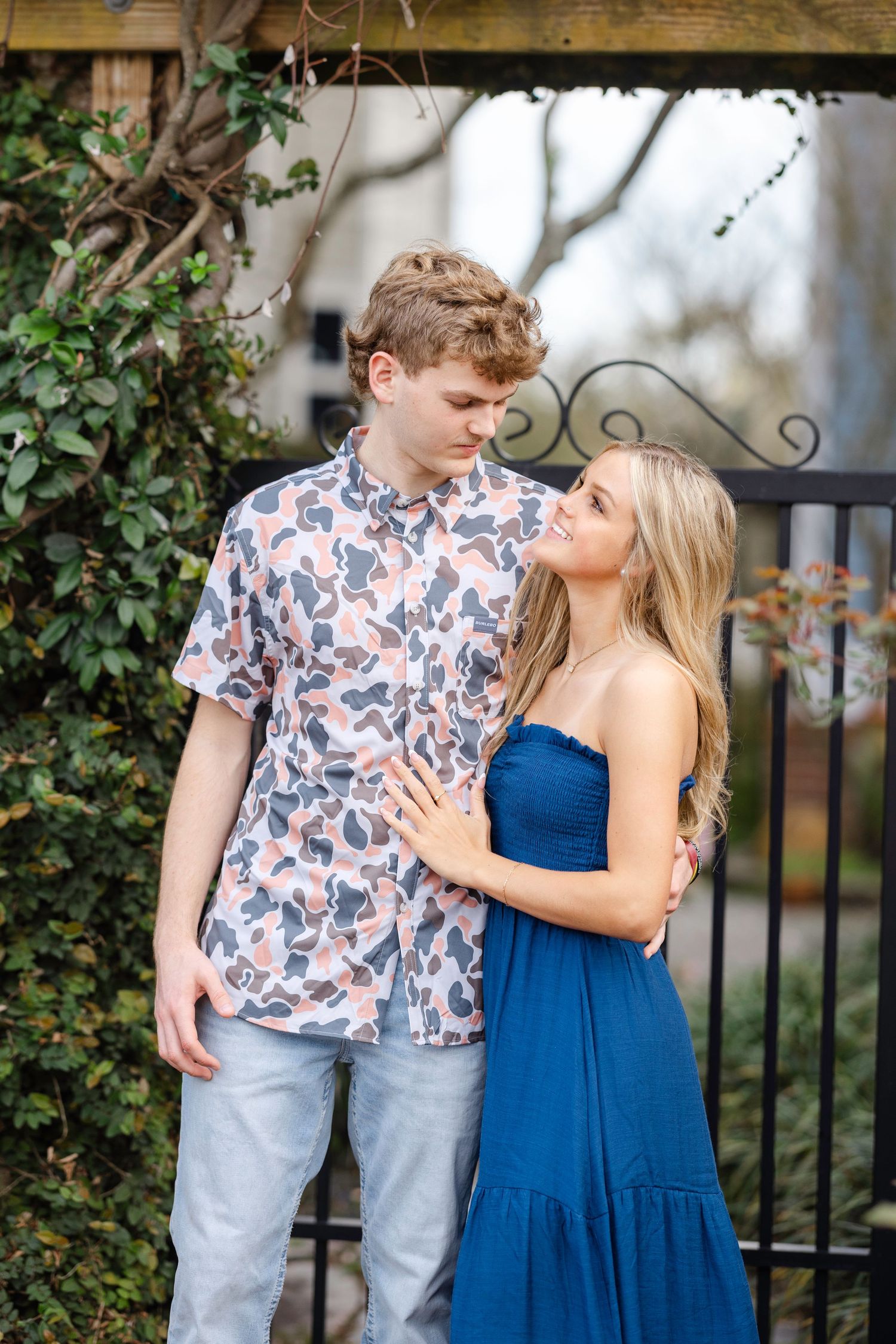 Couple in coordinating blue dress and floral shirt pose together against an ivy-covered wall.