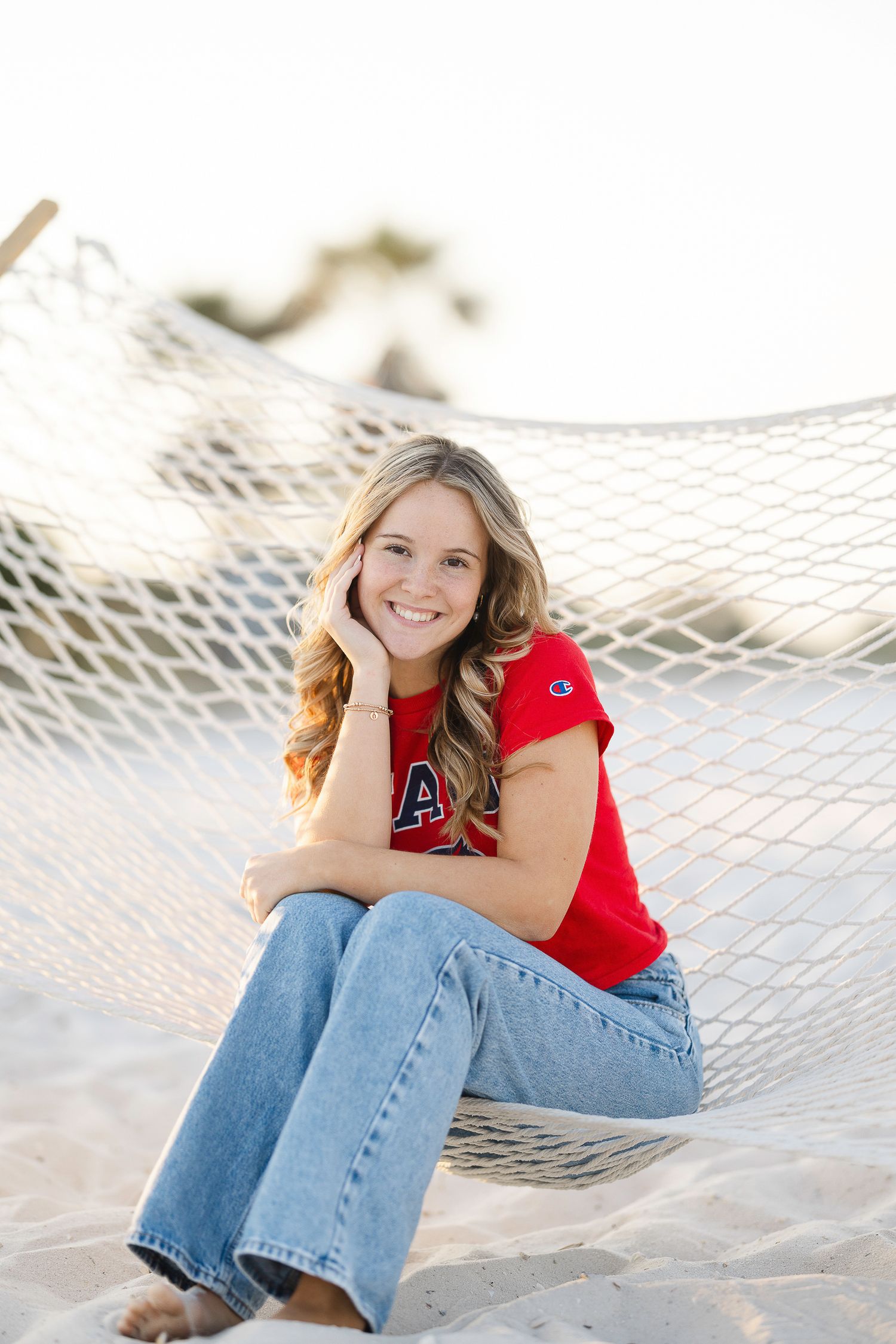 A person in a red top and jeans sits casually on a white hammock at the beach during golden hour.